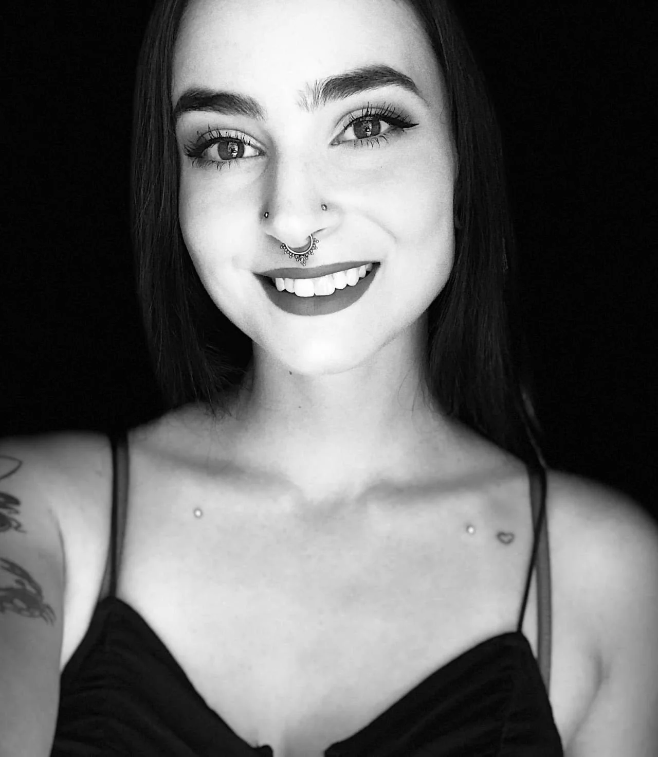 Black and white portrait of a young woman smiling, with piercings, tattooed shoulder, and long dark hair, wearing a spaghetti strap top.
