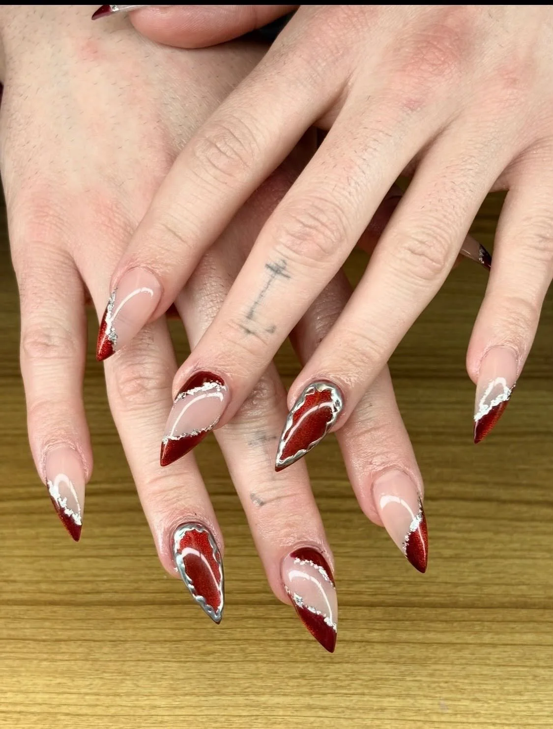 Hands with long, pointed acrylic nails featuring a red and white marble design with silver accents, placed on a wooden surface.
