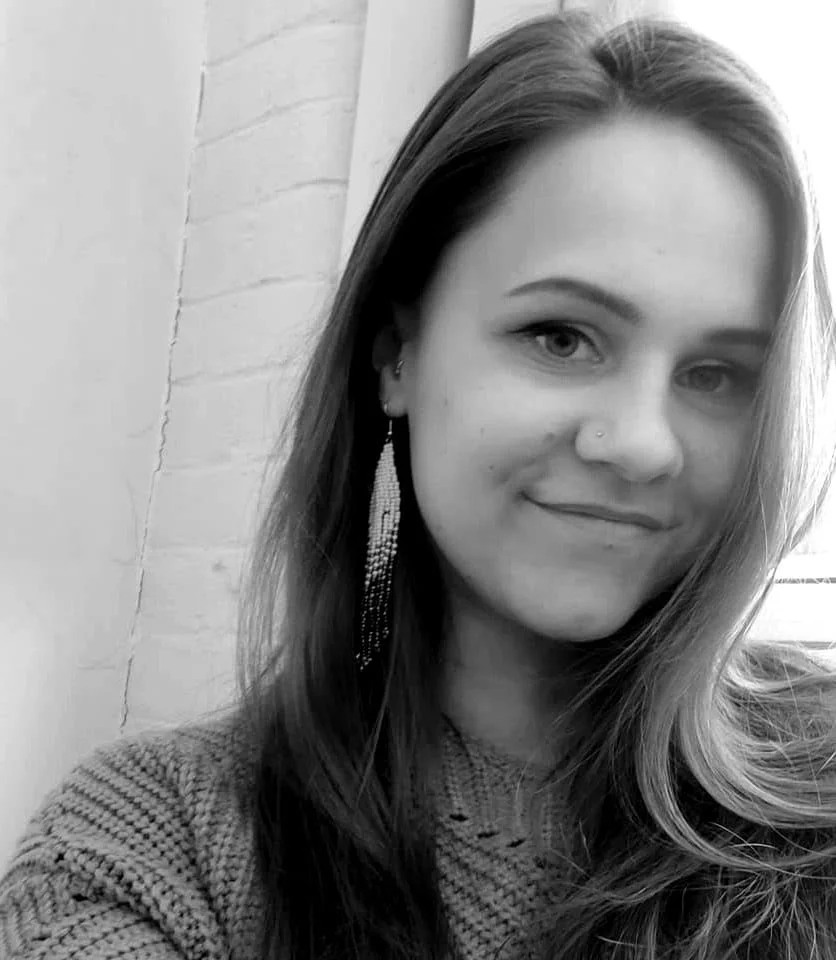 A young woman with long hair and light skin smiling slightly, wearing a textured sweater and long dangling earrings, standing next to a brick wall.