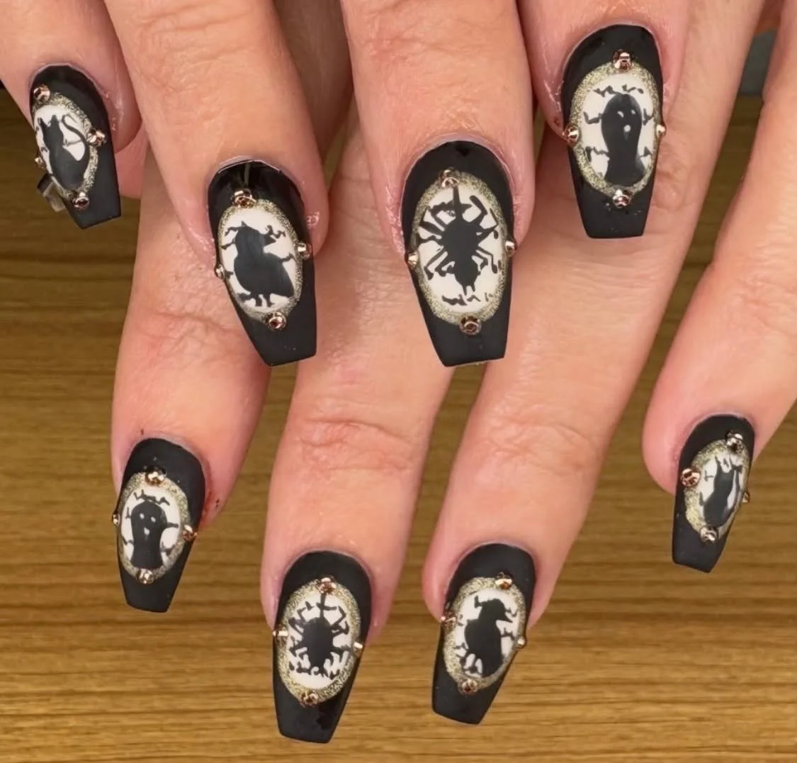 Nail art with black matte base and oval white designs featuring black silhouettes of insects, decorated with small gold and red rhinestones.