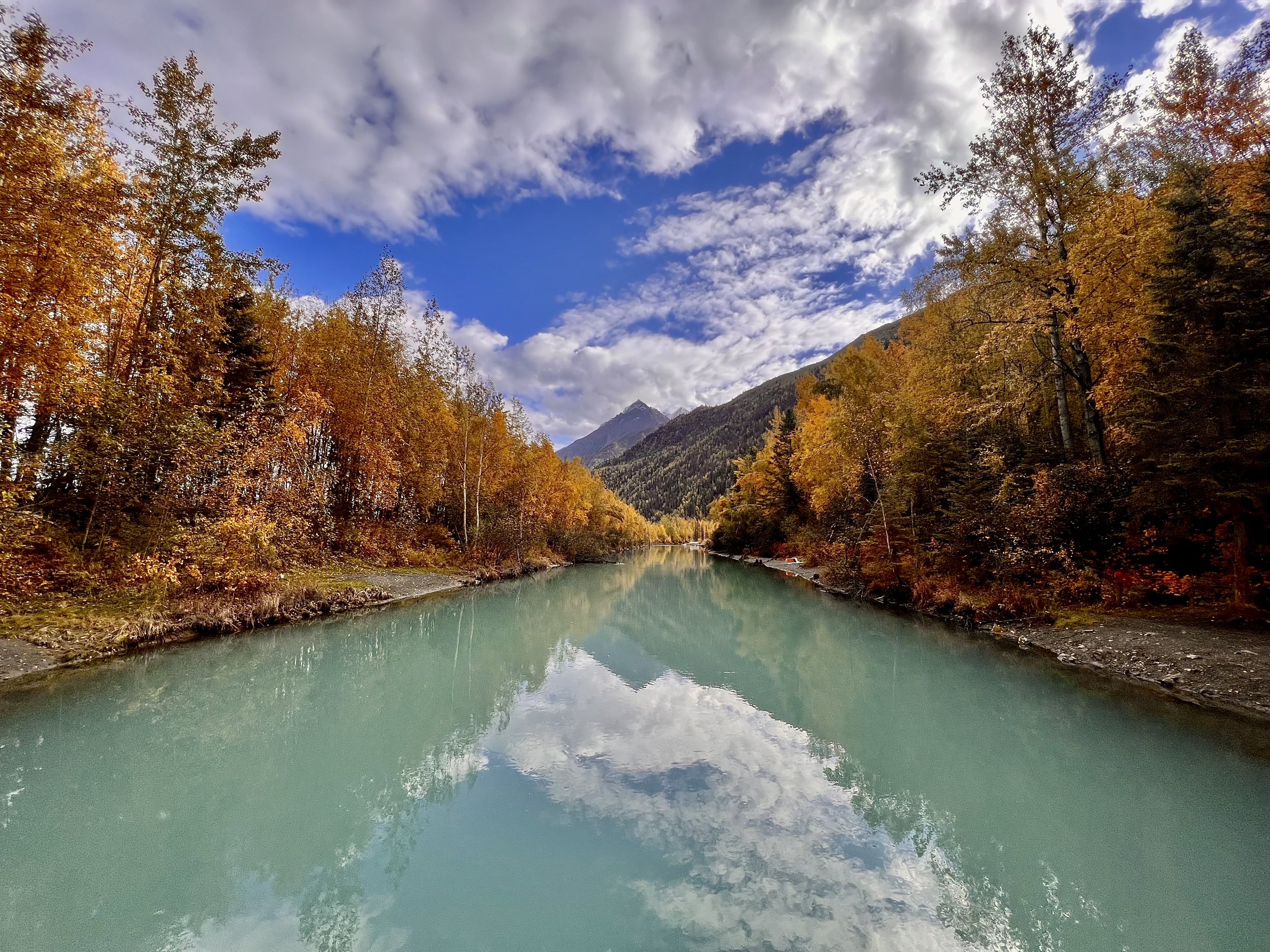 A river flowing through a forest with trees showing fall foliage under a partly cloudy sky with some mountains in the distance.