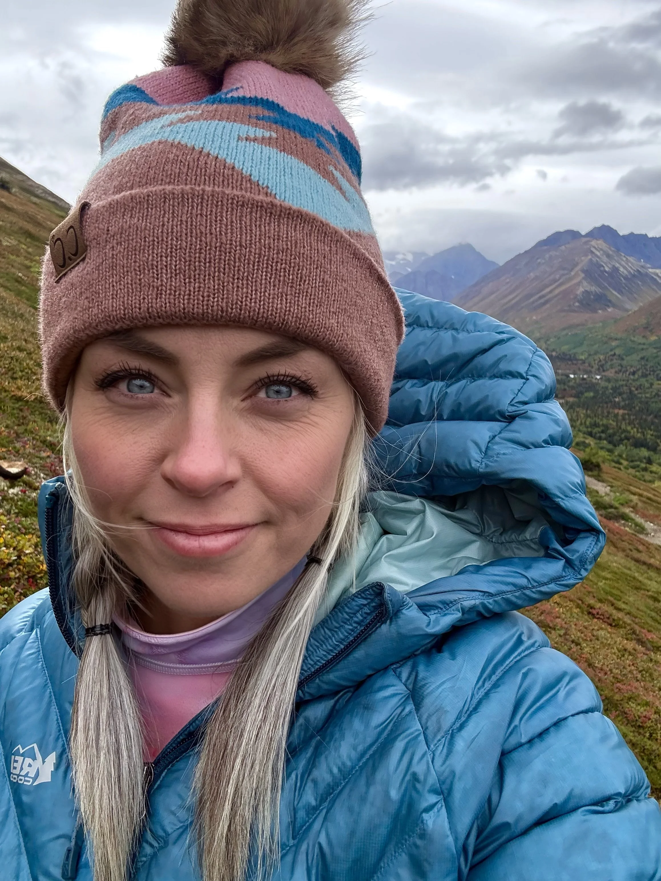 A woman with gray hair and blue eyes taking a selfie outdoors during daytime, wearing a pink beanie with blue stripes, a blue puffy jacket, and a pink shirt underneath. The background features mountains, clouds, and rolling green hills.