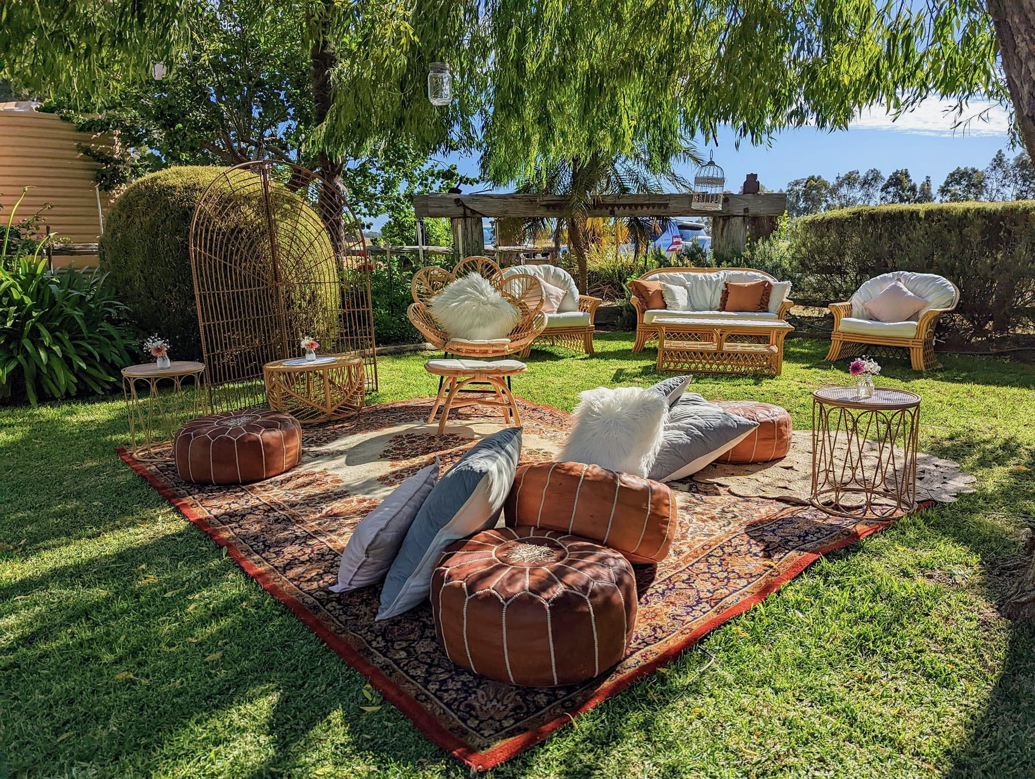 Outdoor patio with wicker furniture, pillows, and a large patterned rug under a tree with hanging mason jars, surrounded by greenery and a fence.