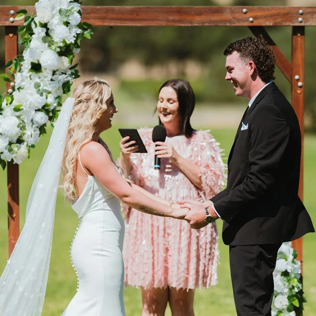 A wedding ceremony outdoors with a bride and groom holding hands, exchanging vows, while an officiant officiates. The scene features a floral arch and a green background.