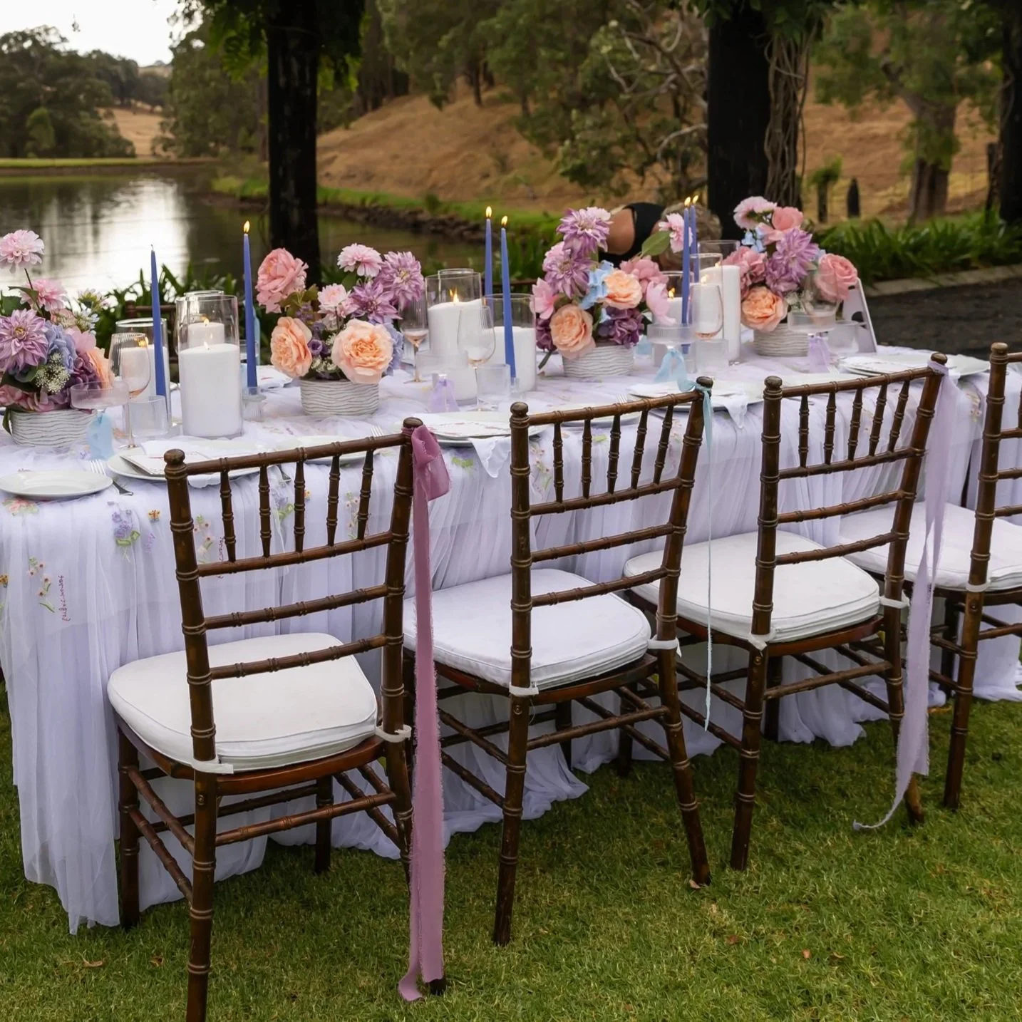 Outdoor wedding or event table set with pink and purple flowers, candles, and tall blue candles, surrounded by wooden chairs with white cushions, near a river with trees in the background.