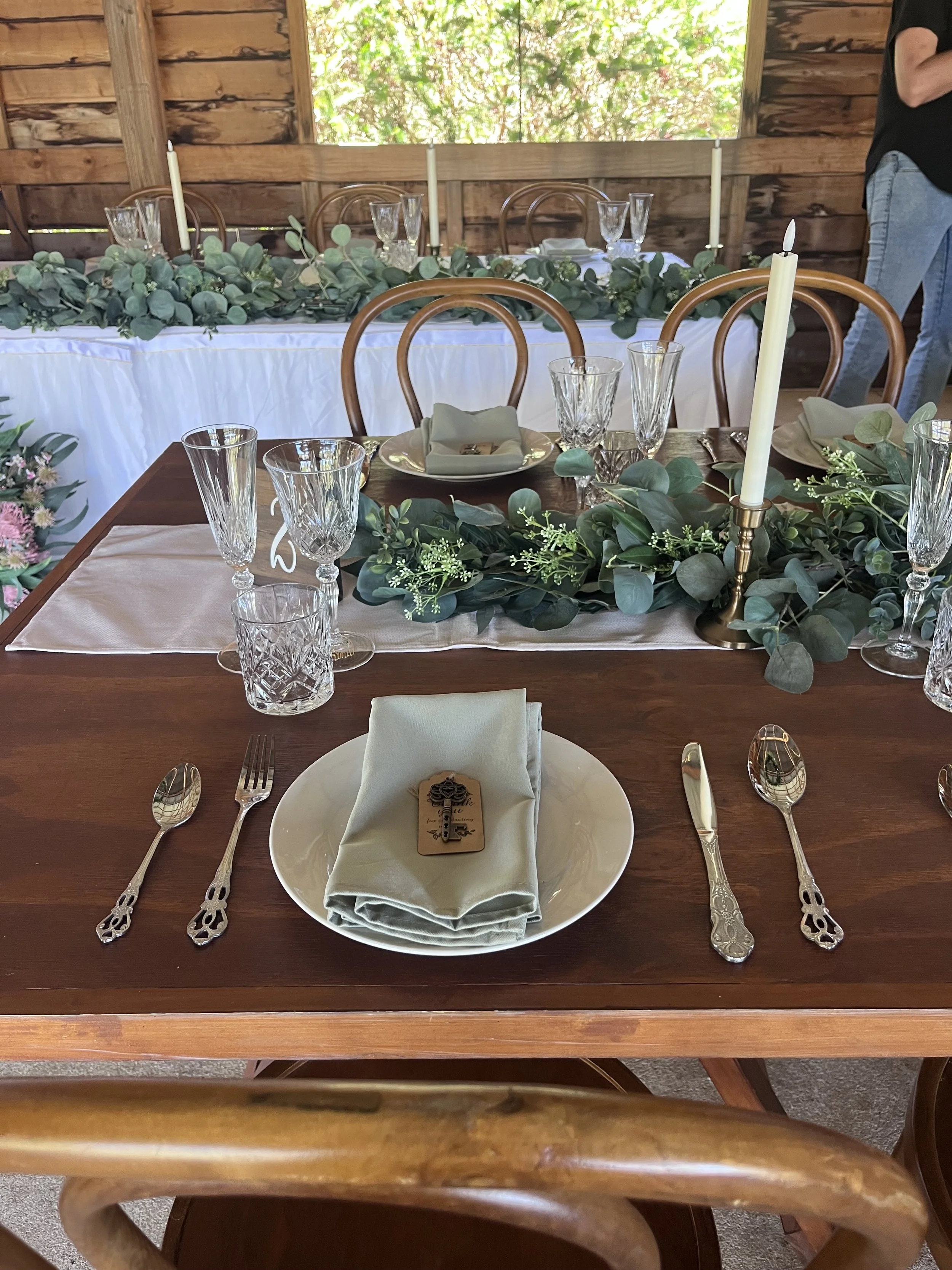 A set dining table with a white tablecloth, surrounded by wooden chairs with curved backs, decorated with green leaves and small white flowers, with no candles. Each place setting includes a white plate, a green cloth napkin, and crystal glassware. A small floral arrangement is on the left side of the table. The background features wooden walls and a window with greenery outside.