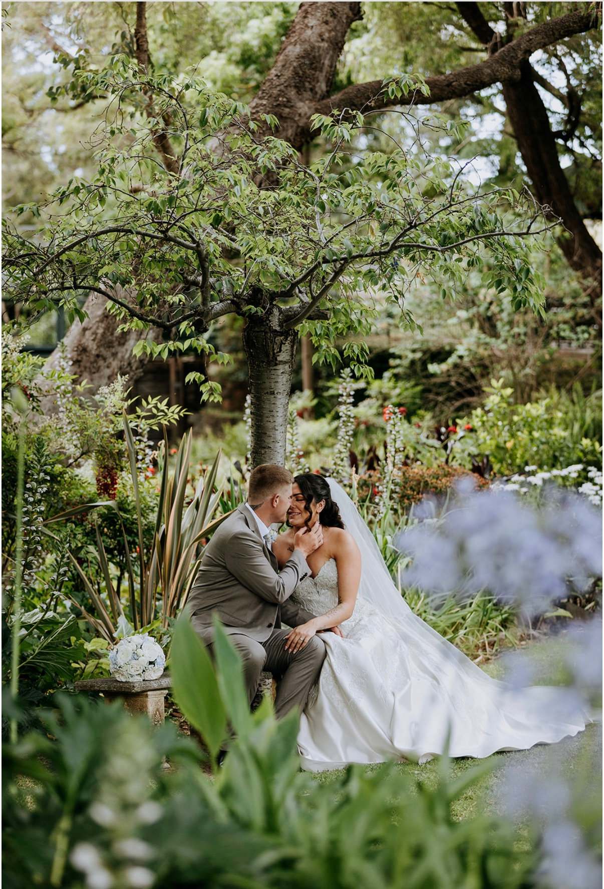 A bride and groom sitting close together in a lush garden, smiling and touching faces under a large tree, surrounded by various green plants and flowers.