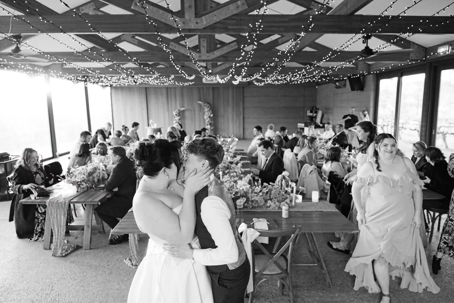 A wedding reception with a bride and groom kissing in the foreground, guests seated at long tables decorated with flowers and candles, string lights hanging from the wooden ceiling, and large windows letting in natural light.