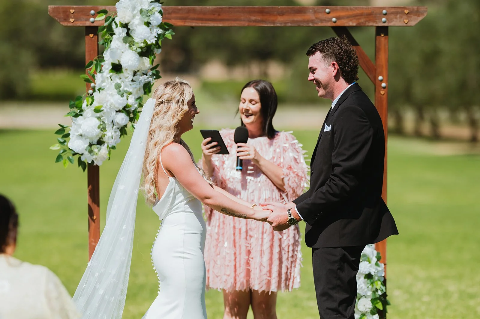 A couple getting married outdoors under a wooden arch decorated with white flowers, with a woman officiating and holding a microphone, on a sunny day with green grass and trees in the background.