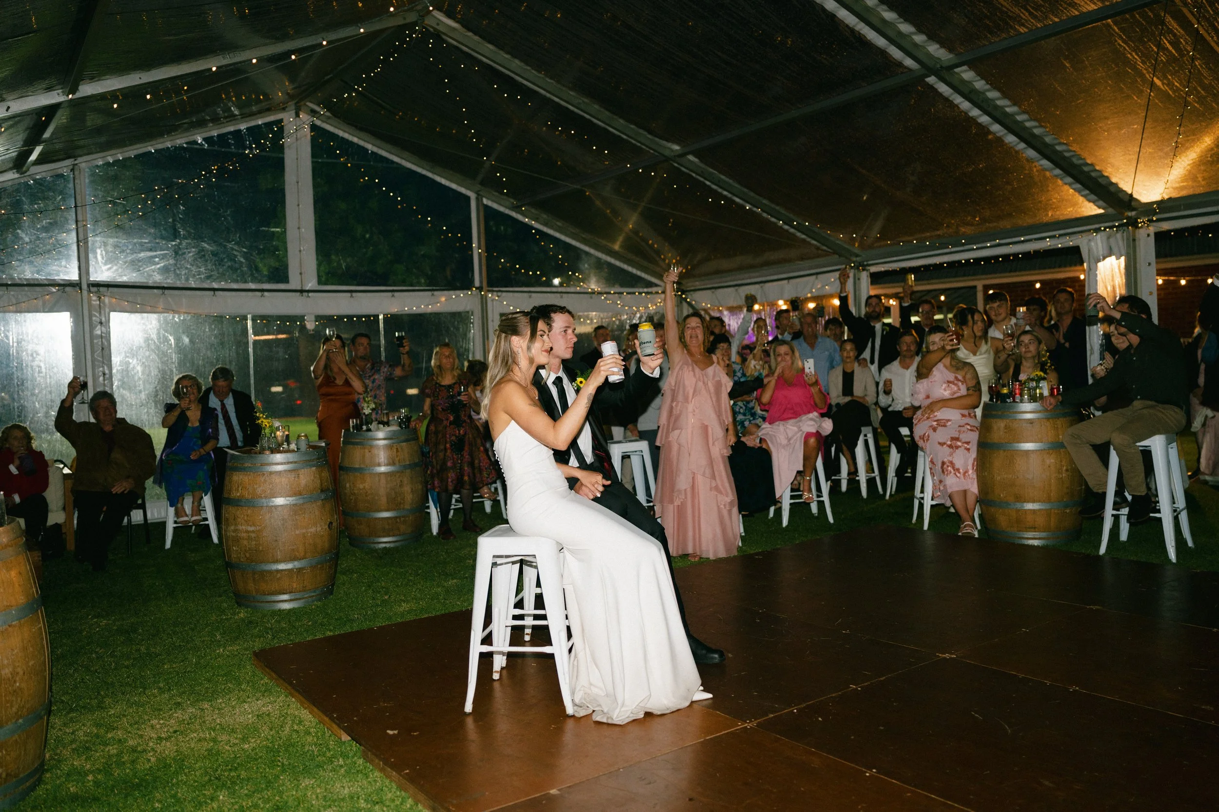 A bride and groom sit on a small stage with a wedding reception in a tent decorated with string lights. Guests are standing and sitting around, some taking photos and raising glasses in celebration.