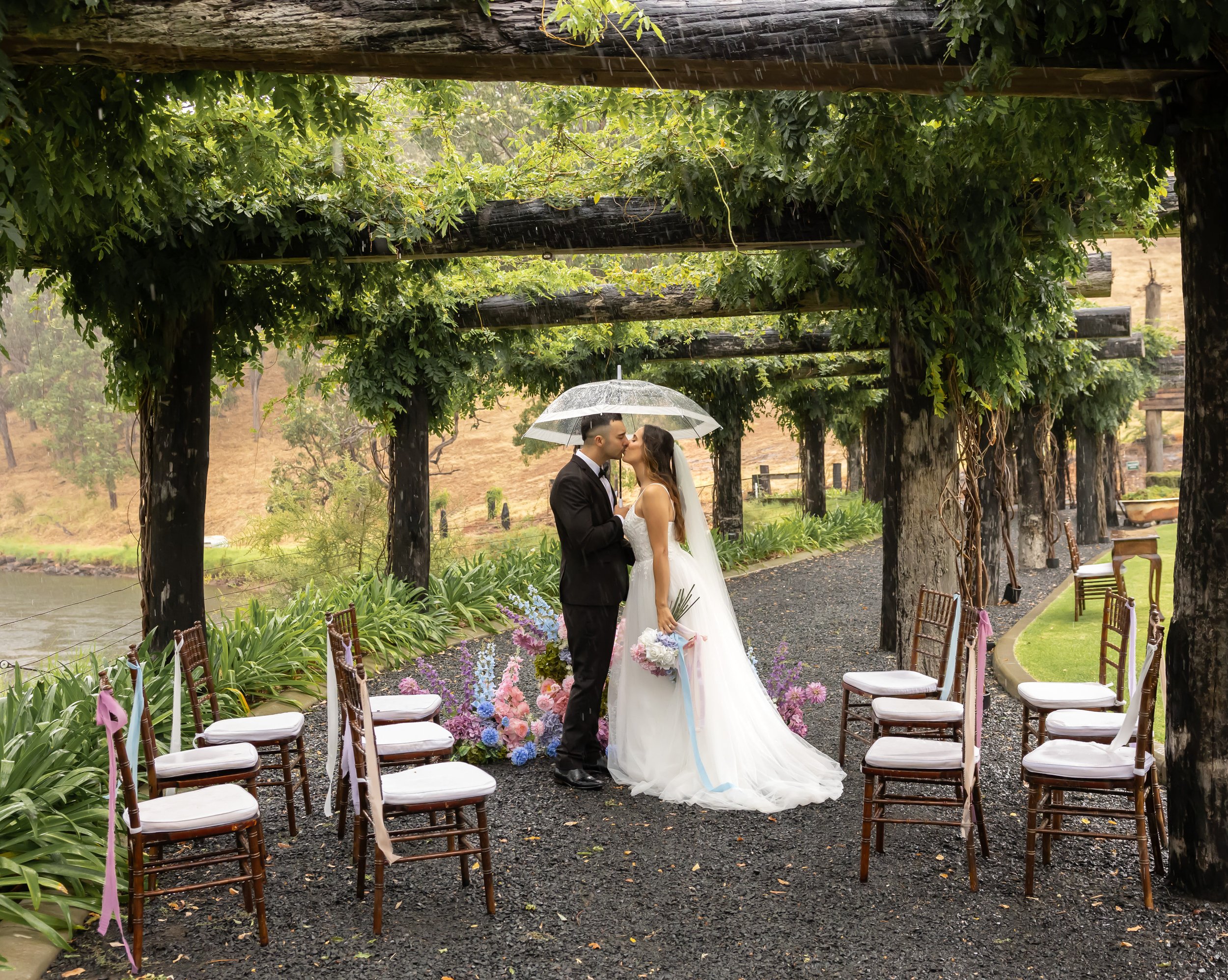 A bride and groom sharing a kiss under a clear umbrella during their outdoor wedding ceremony on a rainy day, with green foliage overhead and chairs arranged for guests.