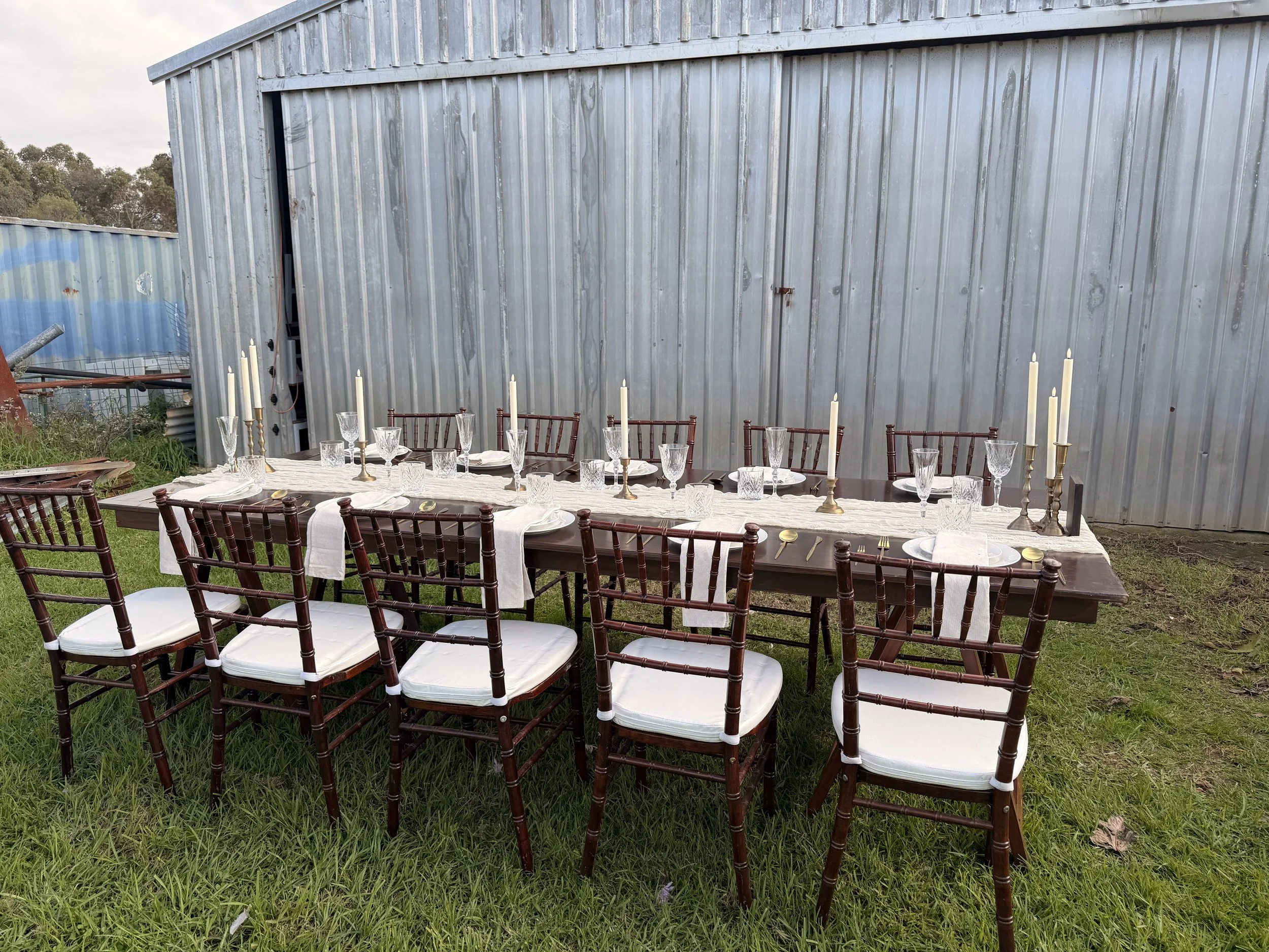 Outdoor dining table set up on grass with chairs, tablecloth, glassware, candles, and cutlery in front of a large gray metal building.