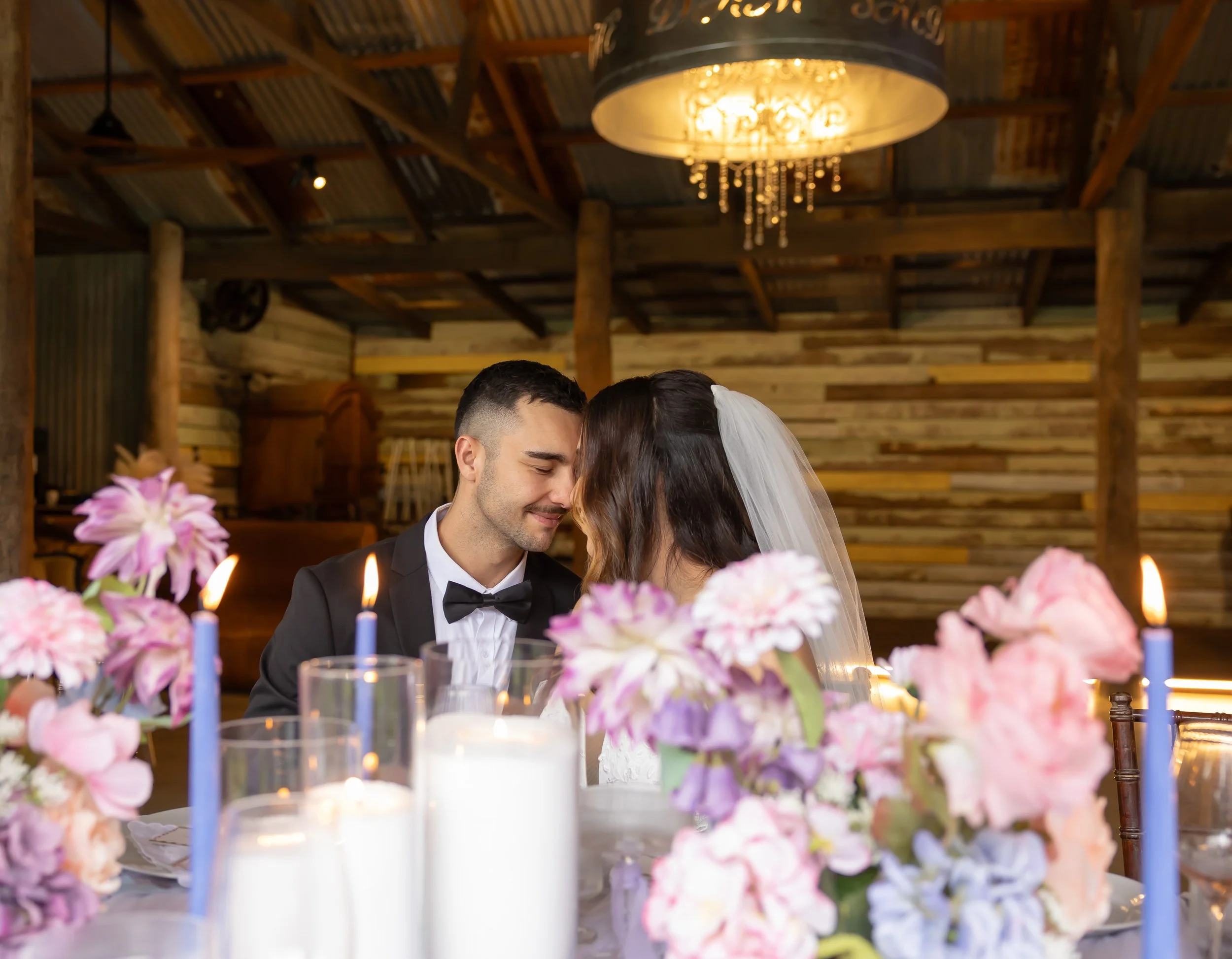 A newlywed couple sharing a kiss at a wedding reception, decorated with pink and purple flowers, candles, and a rustic wooden barn interior.
