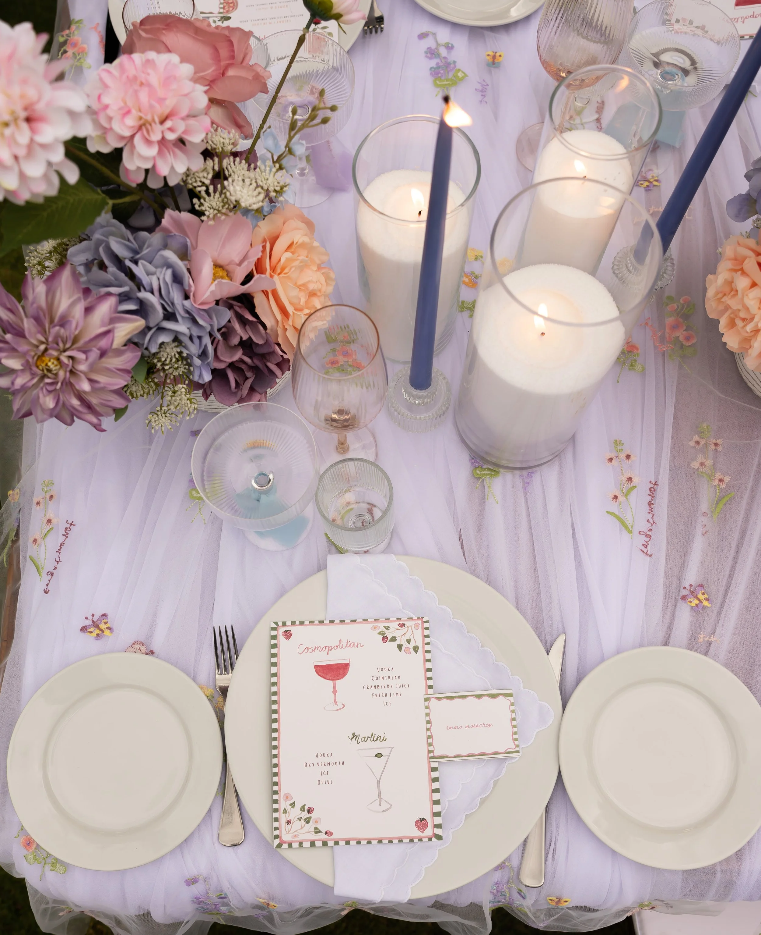 A table set for a celebration with a pink and purple floral centerpiece, white candles in glass holders, and a menu card featuring cocktails such as a cosmopolitan and martini. The tablecloth is white with embroidered details, and there are empty white plates and silverware arranged for guests.