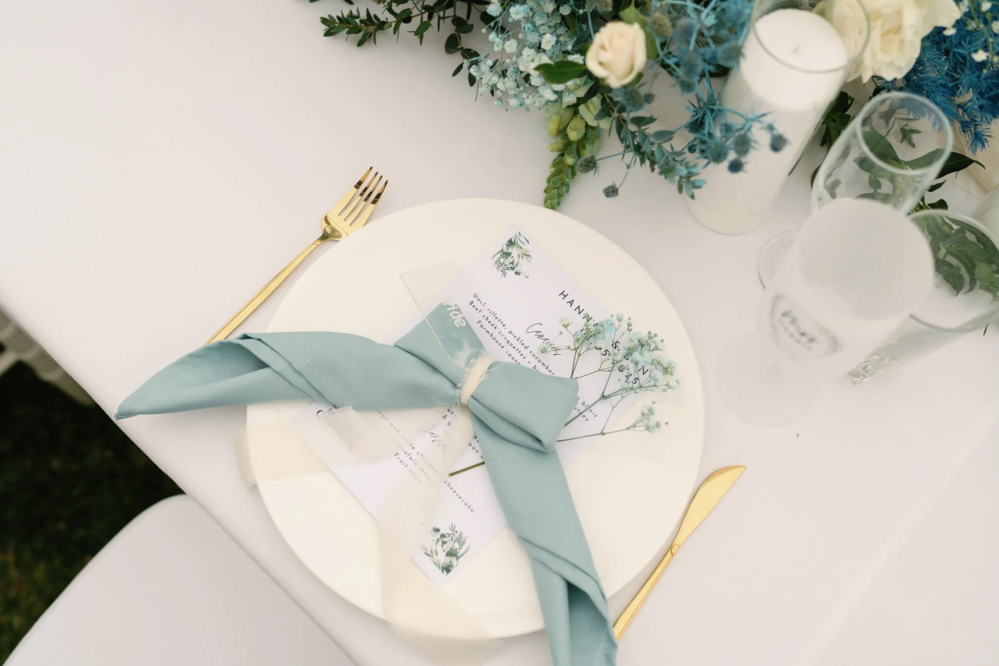 A white round table setting with a gold fork, a folded light blue napkin, and a menu card decorated with botanical illustrations. The table is adorned with a floral centerpiece with white and blue flowers and greenery in white vases.