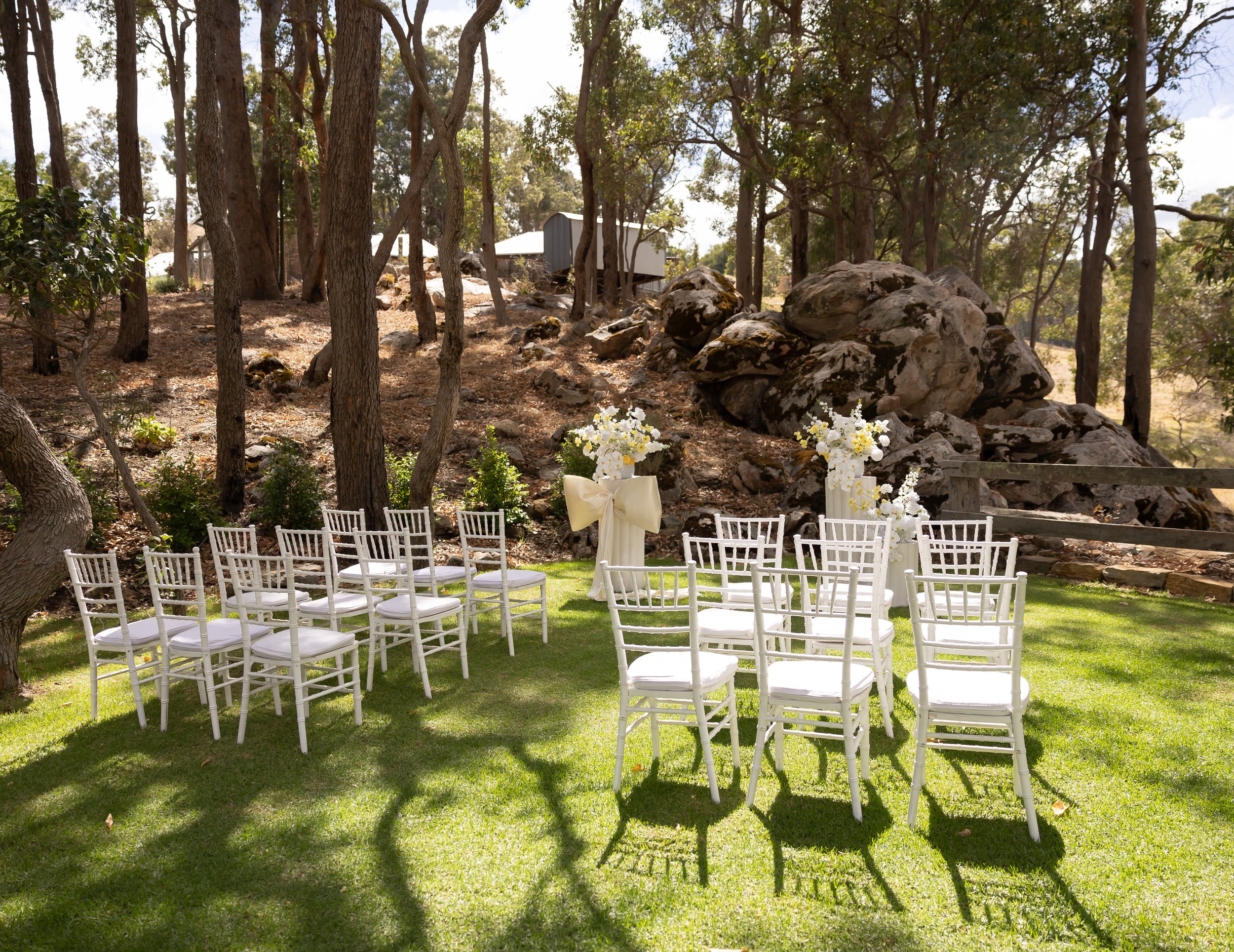 Outdoor wedding setup with white chairs arranged on a green lawn, a tall floral arrangement with white flowers and a yellow bow, surrounded by trees and rocks.