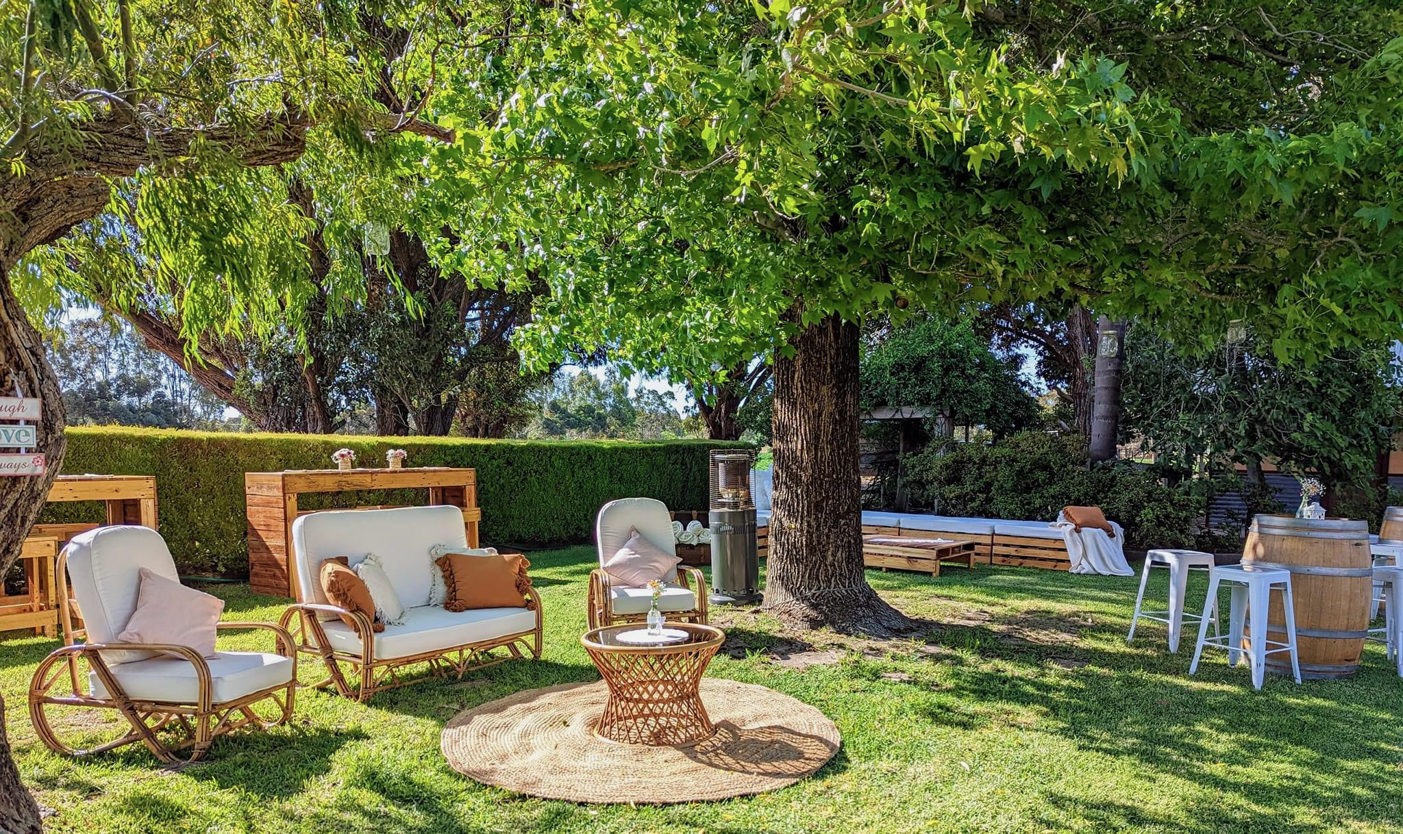 An outdoor patio area with white chairs, a round woven rug, and a wooden coffee table surrounded by trees and greenery. There is a hedge and various potted plants in the background. The setting is sunny and inviting, perfect for relaxing.