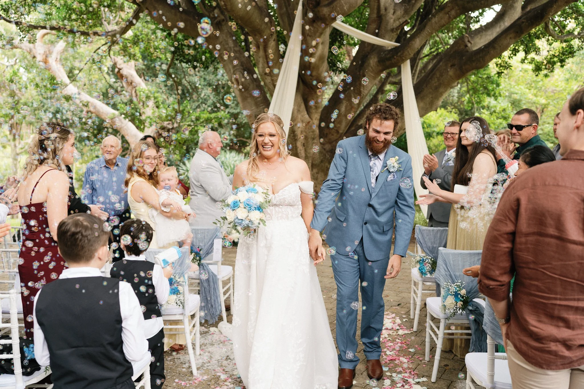 A group of people at a wedding ceremony outdoors, with the bride and groom walking away, smiling. The audience is celebrating by blowing bubbles and clapping, surrounded by green trees and chairs decorated with fabric.