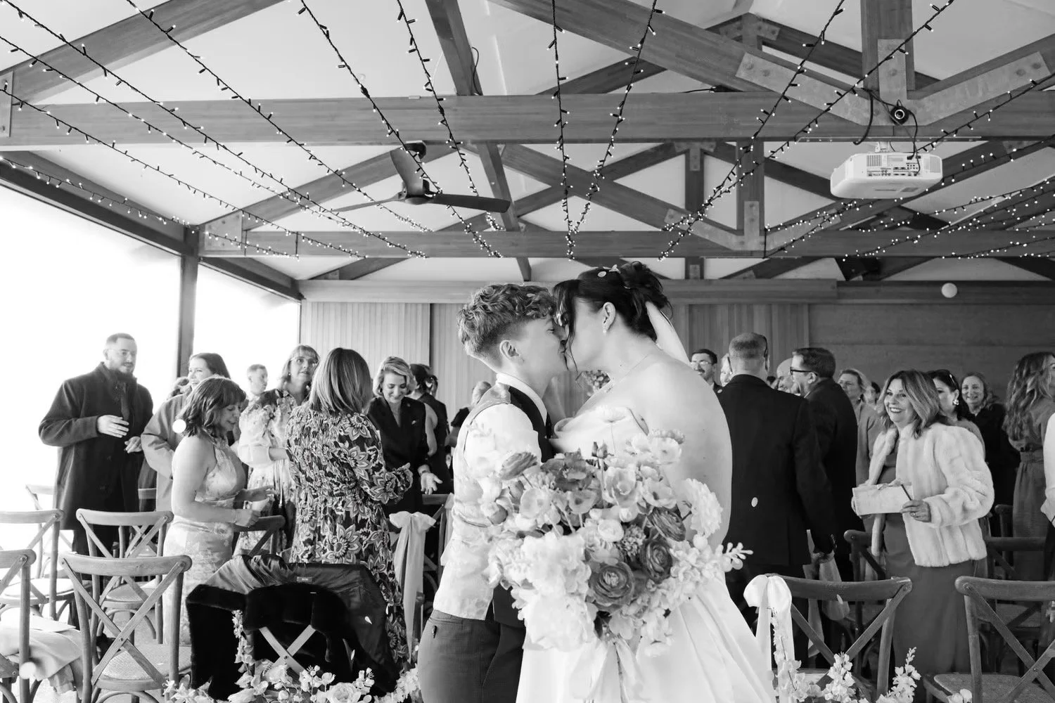 A bride and groom share a kiss at their wedding reception, surrounded by guests. The bride holds a large bouquet of flowers, and the groom wears a vest and tie. The reception hall is decorated with string lights on a wooden ceiling, and there are chairs and people in the background celebrating.