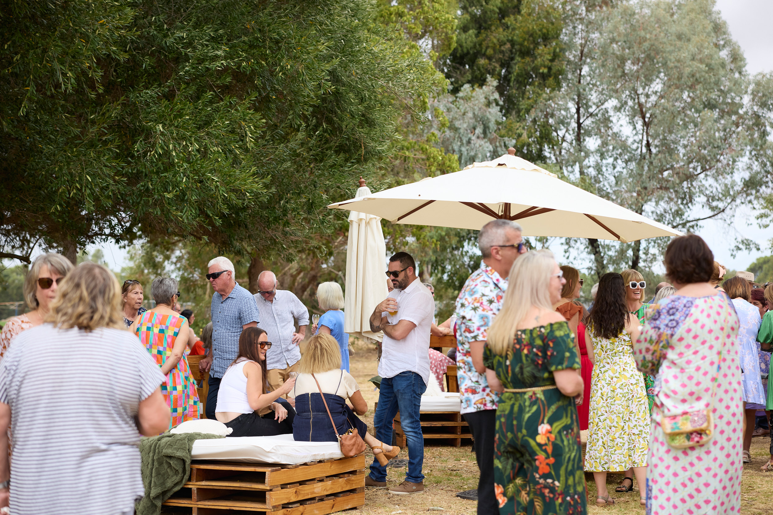 People gather outdoors near large trees and a white umbrella, some wearing sunglasses and colorful clothing, at a sunny event or festival.
