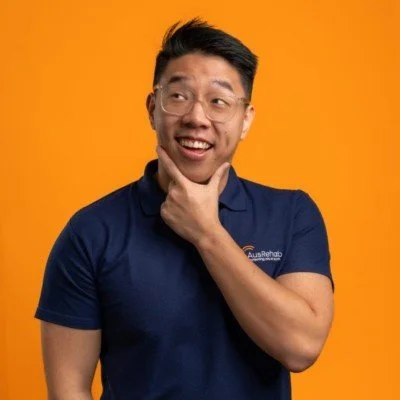 A man with glasses, wearing a blue polo shirt, smiling and thinking with his hand on his chin against an orange background.