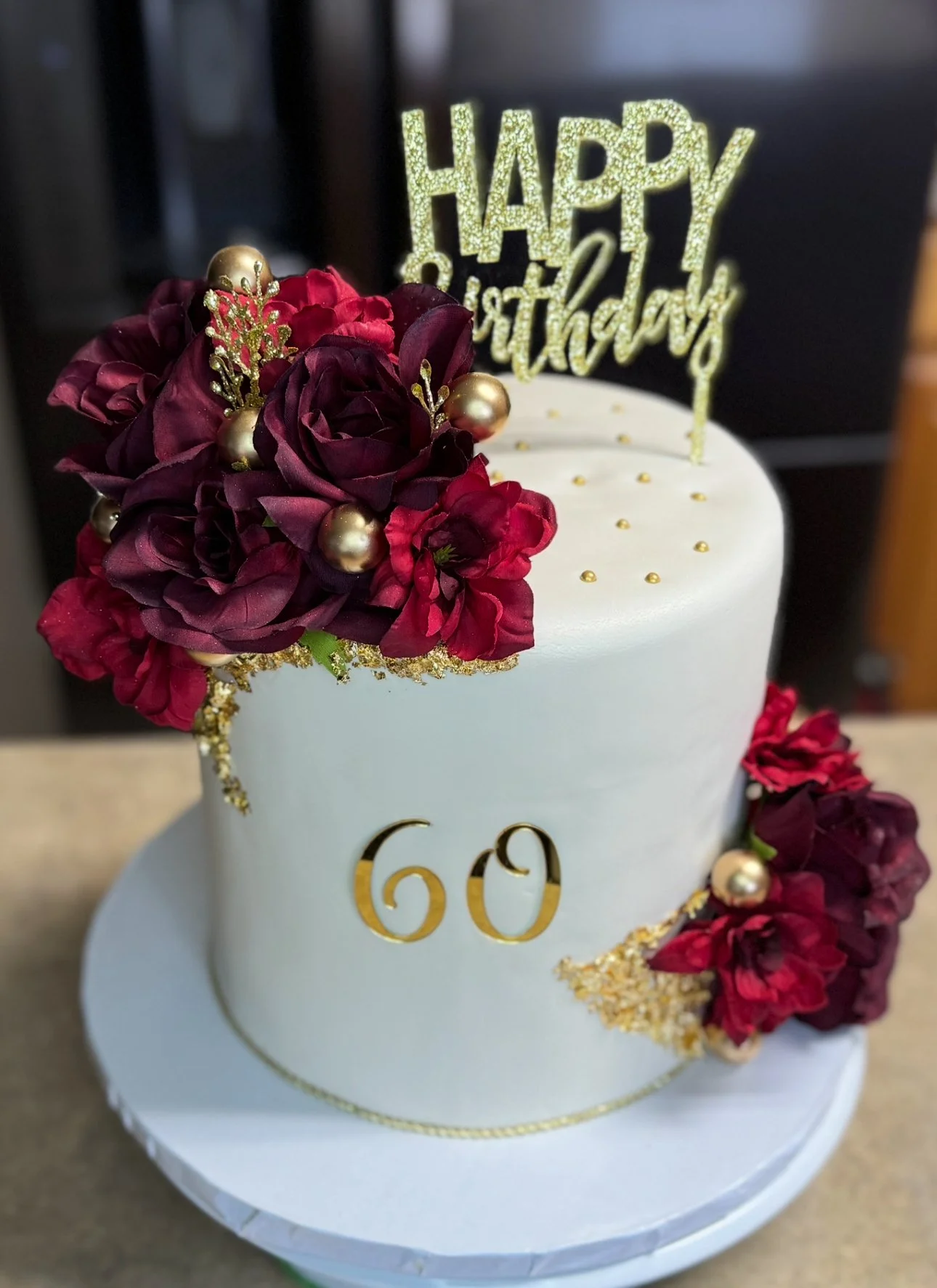 A white birthday cake with gold decorations, a 'Happy Birthday' topper, dark red flowers, gold beads, and the number 60 in gold.