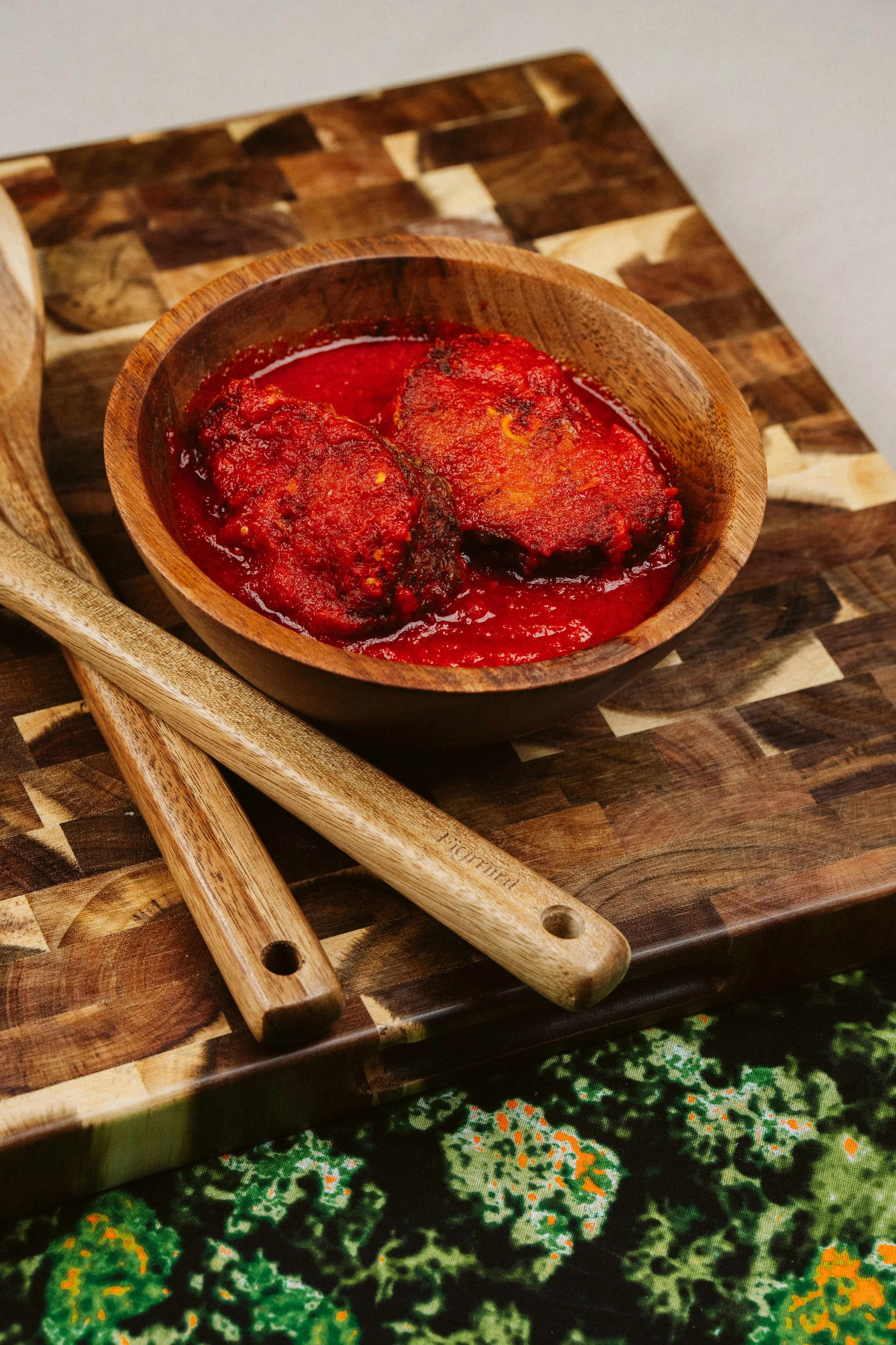 A wooden bowl containing two large meatballs in red sauce, placed on a wooden cutting board with a patterned fabric underneath.