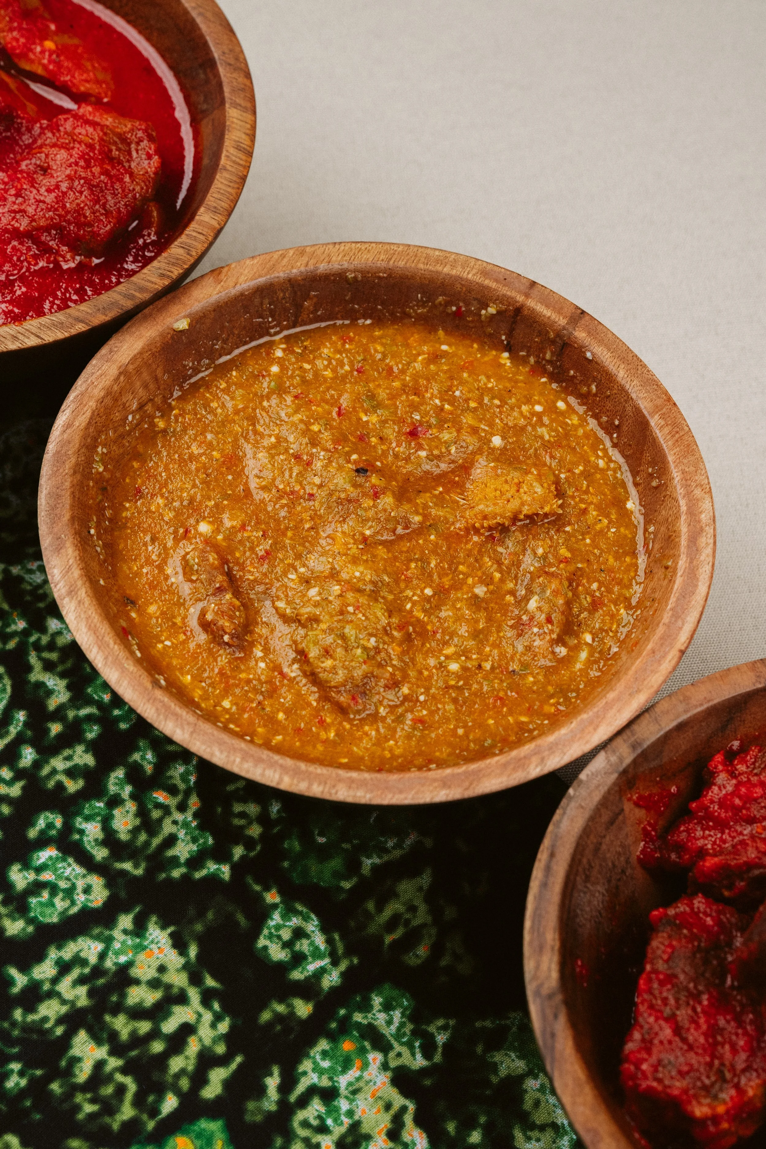 Close-up of two small wooden bowls containing different types of chili sauces or salsas, with one in focus showing a chunky yellow-orange sauce and the other partially visible with a red sauce, placed on a patterned green tablecloth.