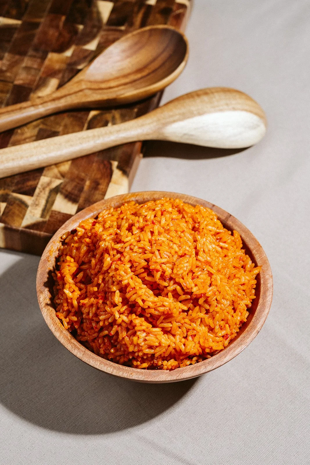 A wooden bowl filled with spicy orange-colored rice on a gray surface, with two wooden spoons and a wooden cutting board in the background.