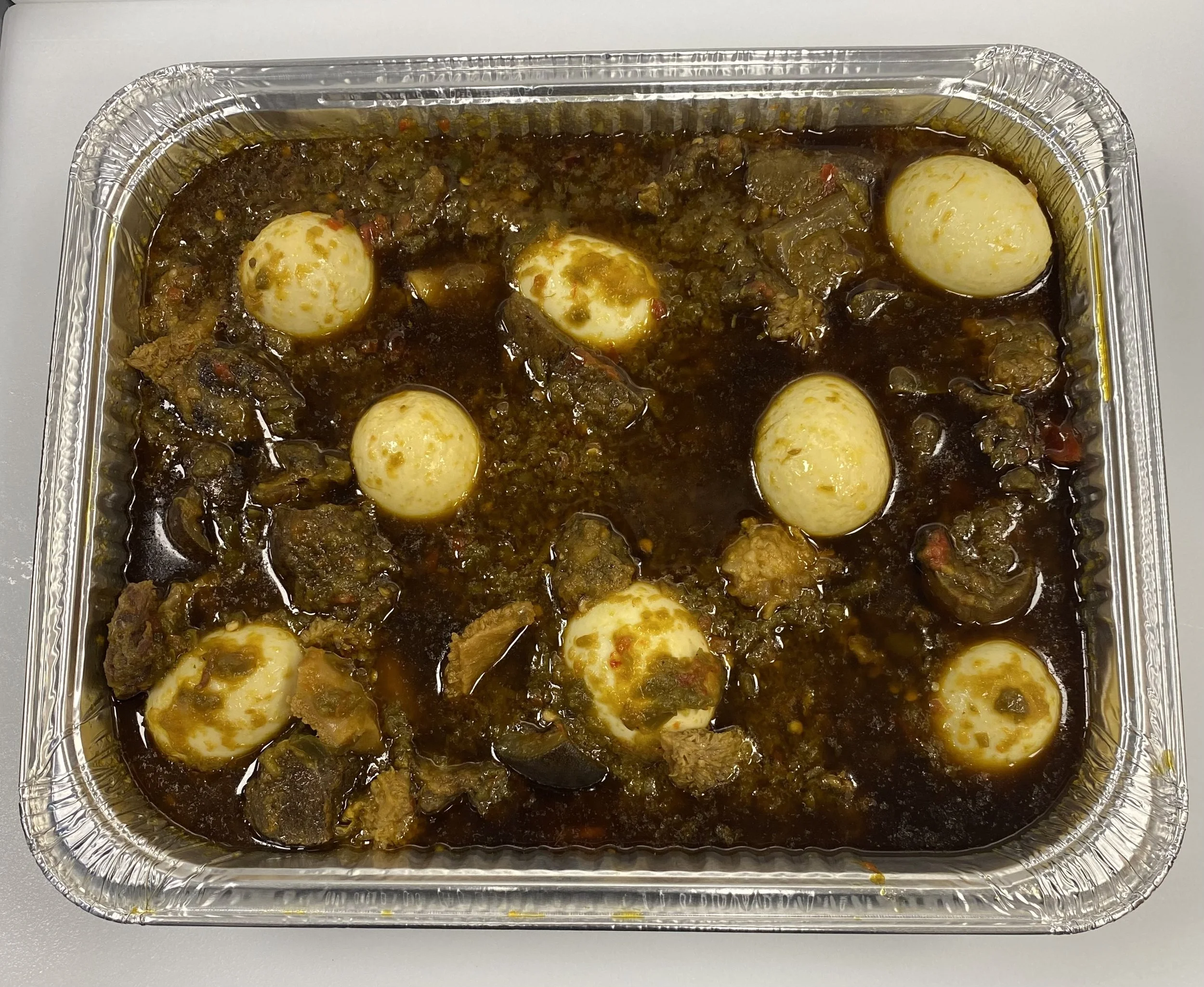 A aluminum tray filled with a dark cooked stew containing eggs, vegetables, and meat.