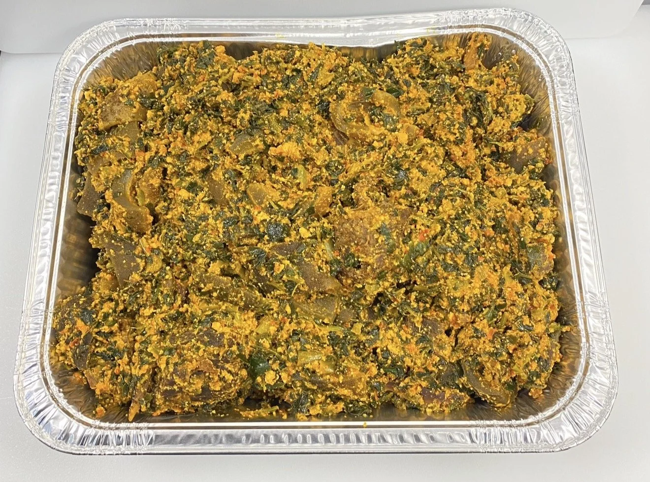 Tray of cooked collard greens with a yellow seasoning or sauce, possibly containing spices and chopped vegetables.
