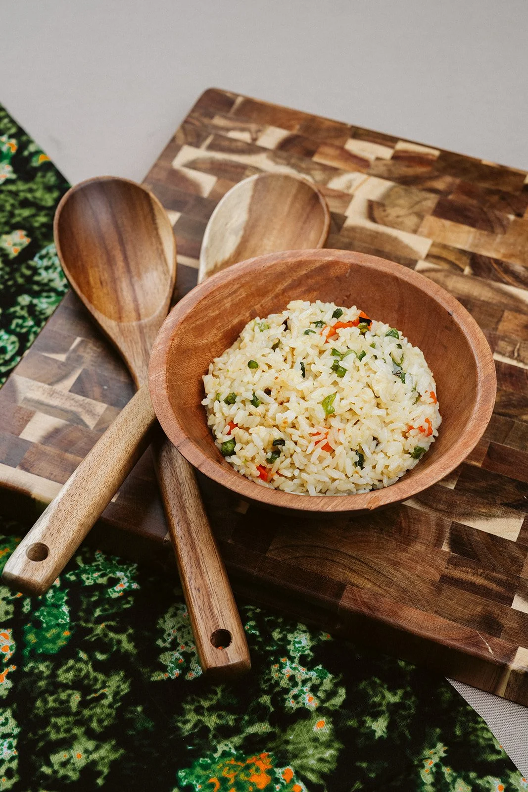 A wooden bowl filled with fried rice and vegetables on a wooden cutting board, accompanied by two wooden spoons.