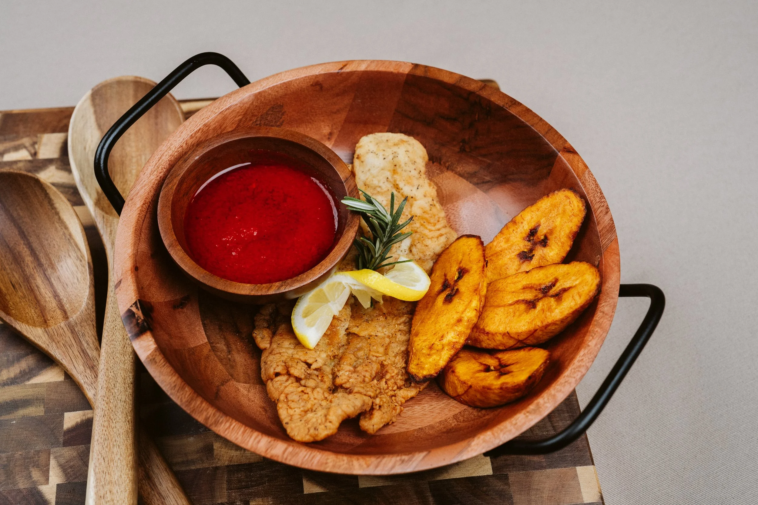 A wooden tray filled with fried fish, fried plantains, lemon slices, potato wedges, and a small bowl of red sauce garnished with rosemary.