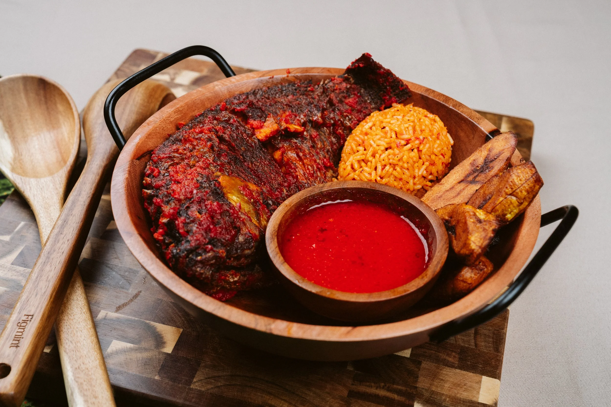 A wooden bowl with a grilled fish covered in red sauce, a serving of orange rice, a small bowl of red sauce, and plantains, on a wooden table.