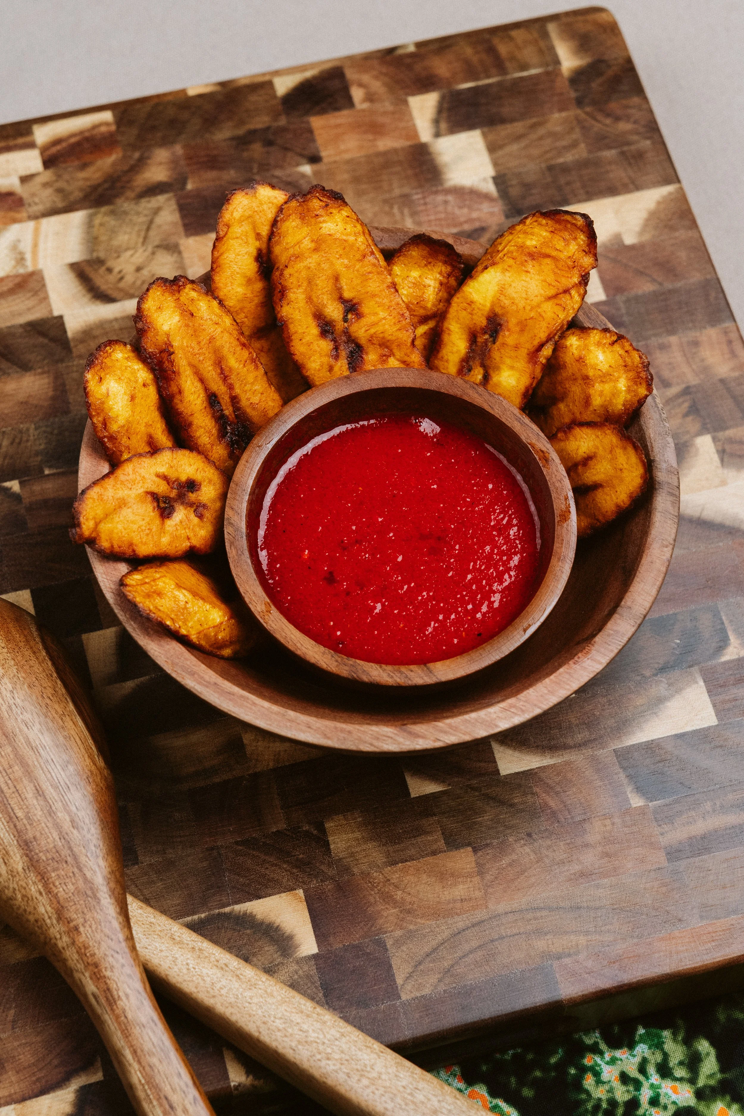 Fried plantains served with a side of red dipping sauce on a wooden platter with wooden utensils and a background of a wooden table.