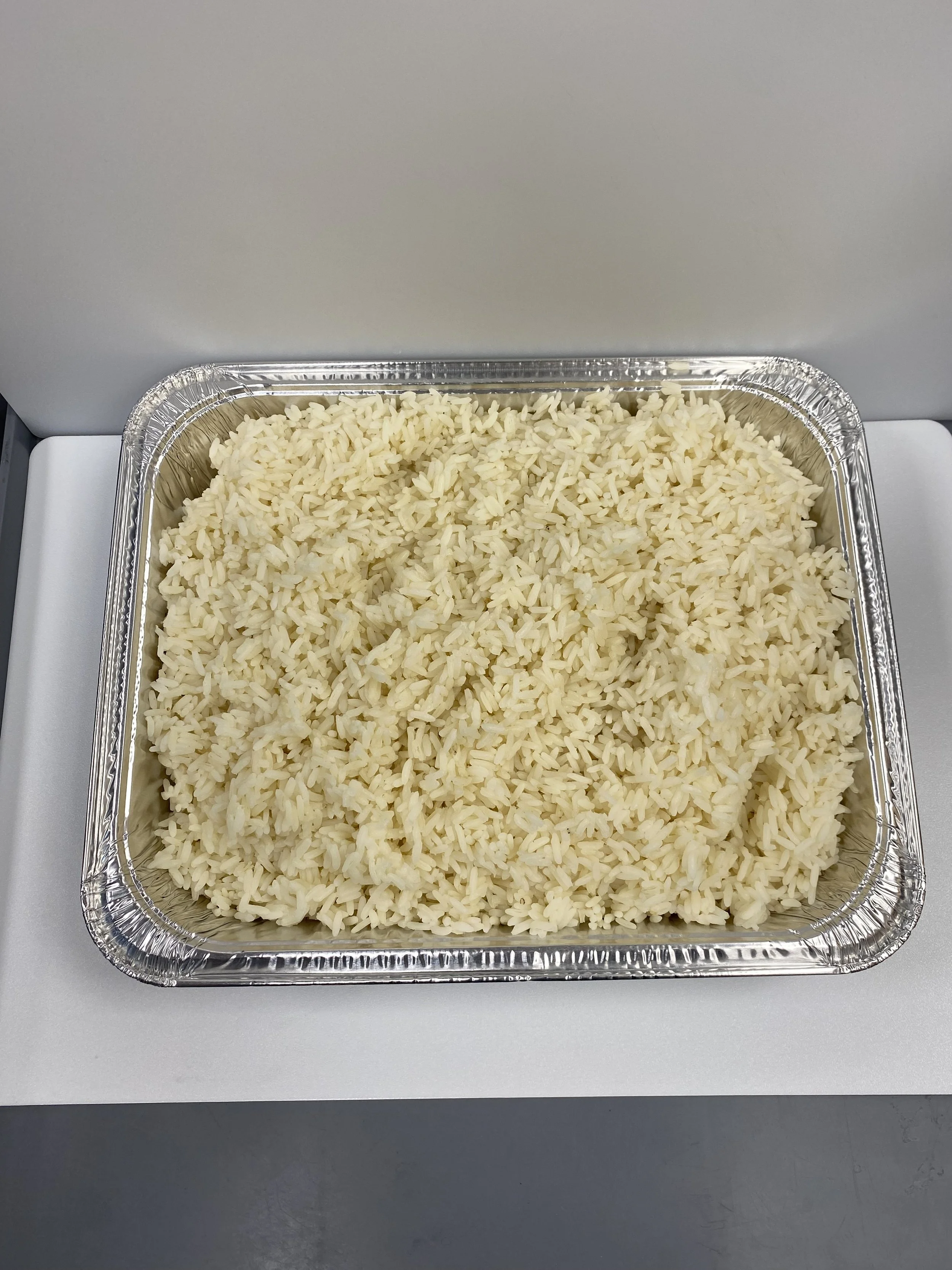 Aluminum tray filled with cooked white rice on a white surface, with a plain gray background.