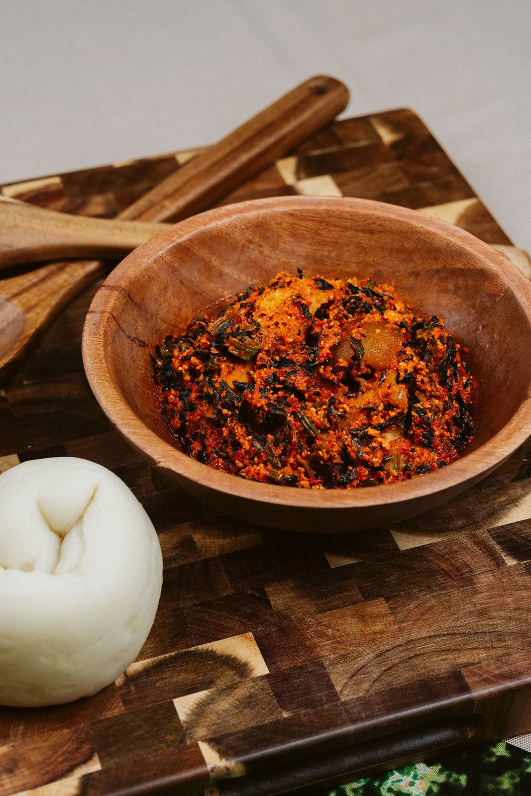 A wooden bowl containing a spicy, cooked vegetable dish with dark green leaves and orange-red sauce, placed on a cutting board with a piece of bread and a wooden spoon nearby.