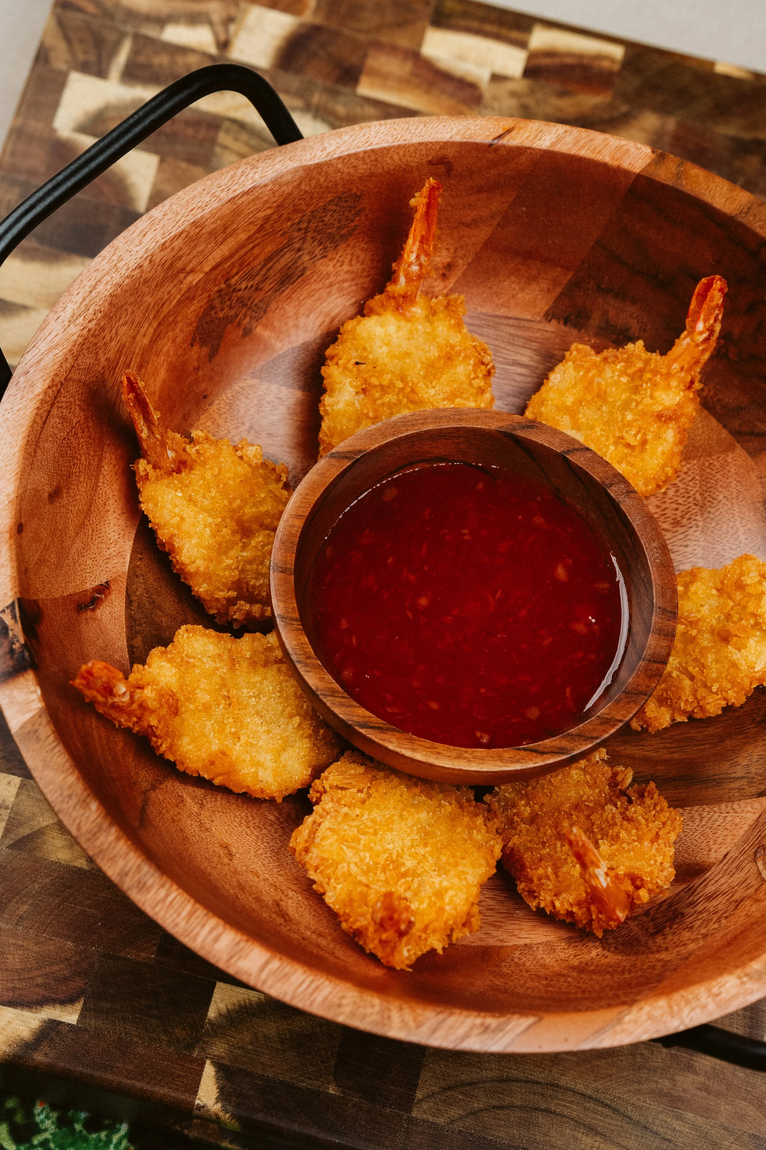 A wooden bowl containing several pieces of fried shrimp with tails, served with a small cup of red dipping sauce.