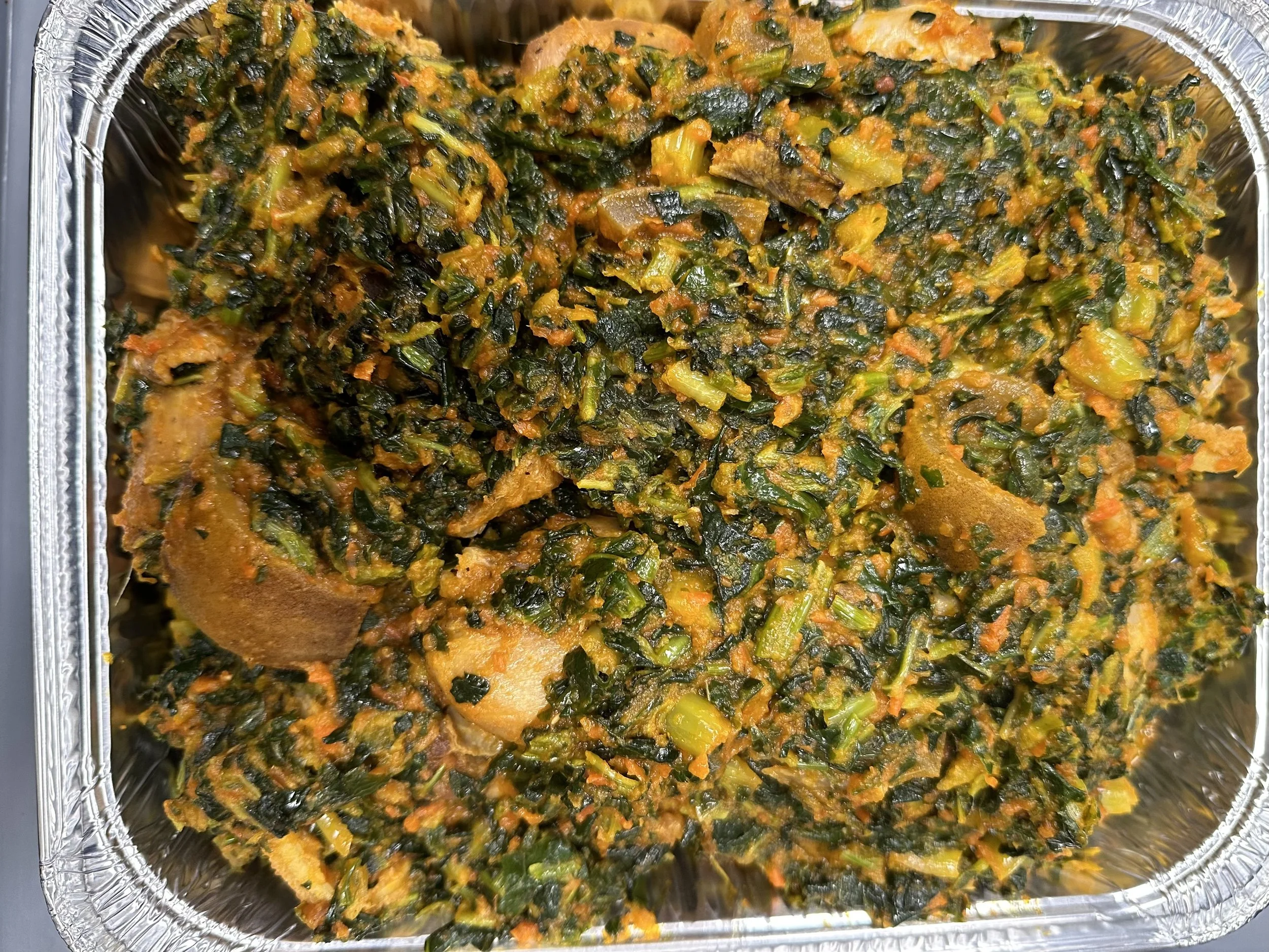 A close-up view of a foil container filled with cooked spinach and other greens mixed with orange-colored seasoning or sauce.