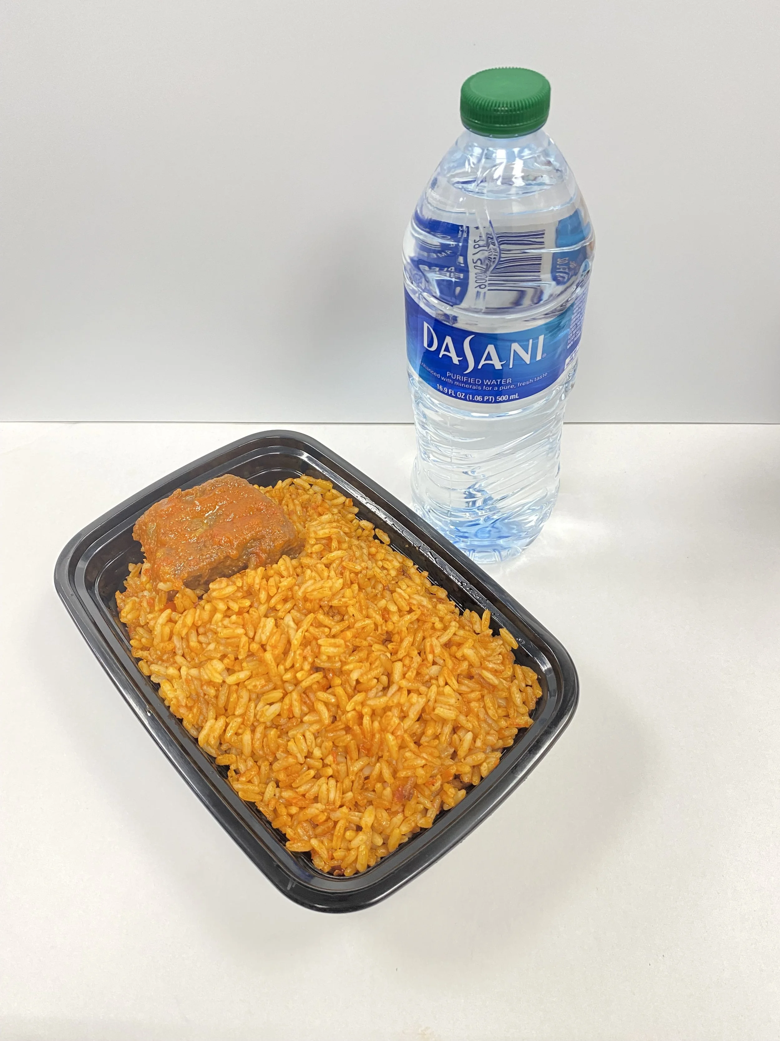 A black plastic tray of rice covered in tomato sauce, with a meat patty on top, next to a bottle of Dasani purified water with a green cap, on a white table against a plain background.