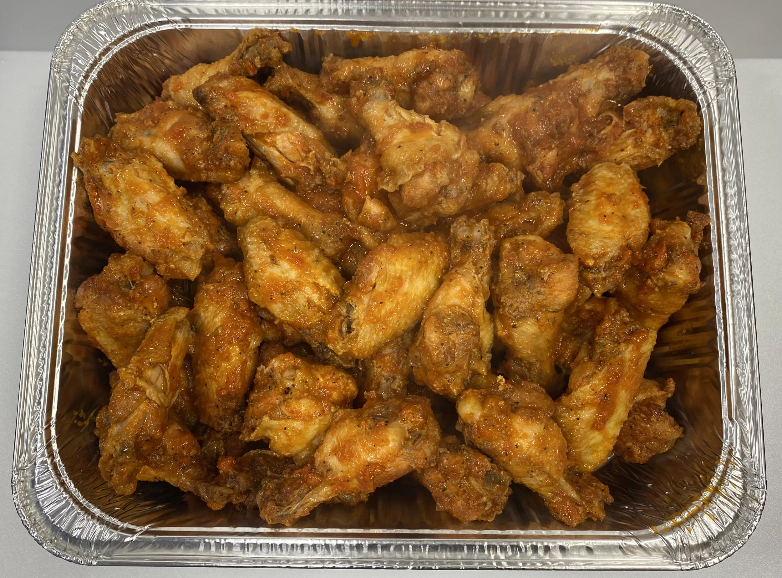 Aluminum takeout container filled with cooked chicken wings coated in spicy sauce.