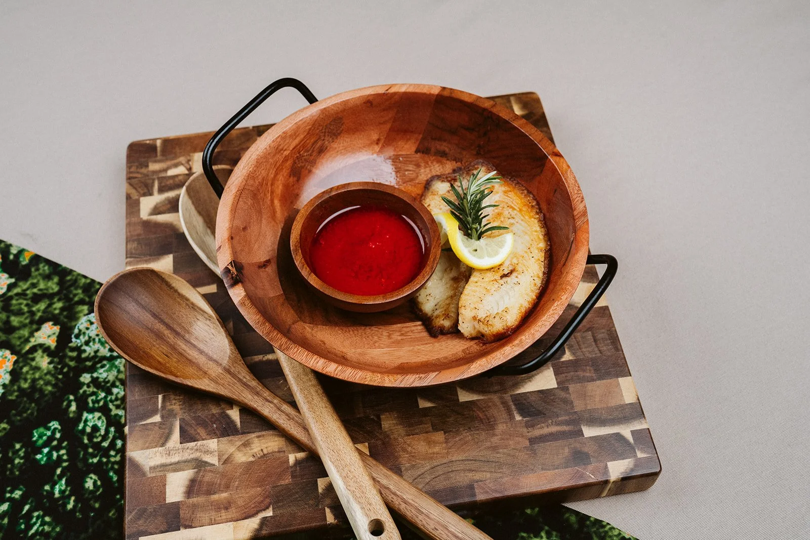 A wooden bowl with a lemon slice and rosemary on a piece of fish, with a small cup of red sauce, placed on a wooden tray with two wooden spoons beside it.
