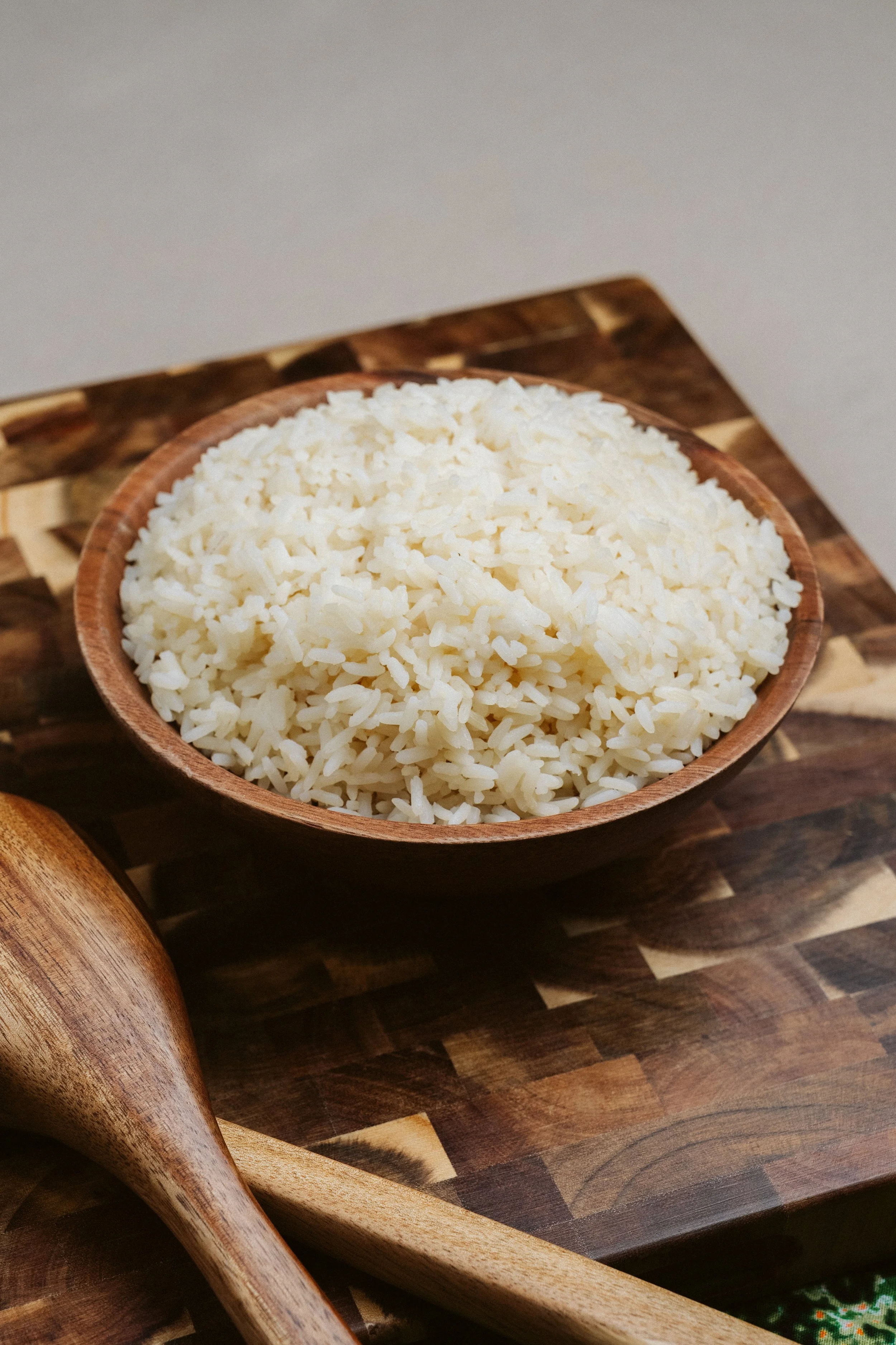 A bowl of cooked white rice on a wooden cutting board, with a wooden spoon nearby.
