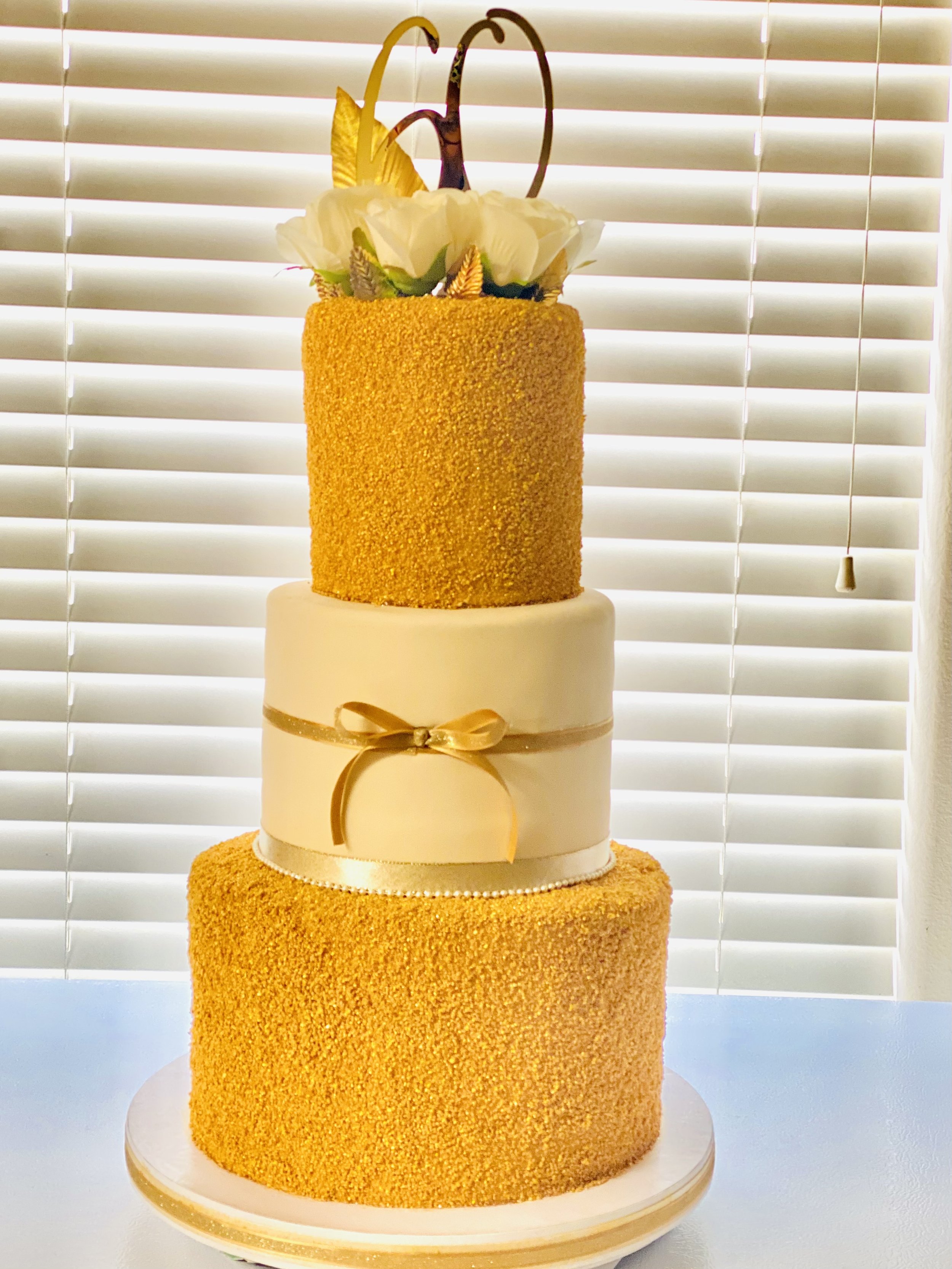 A three-tiered wedding cake with a gold and white color scheme, decorated with gold ribbon and a white flower arrangement at the top, placed on a white cake stand in front of closed white window blinds.