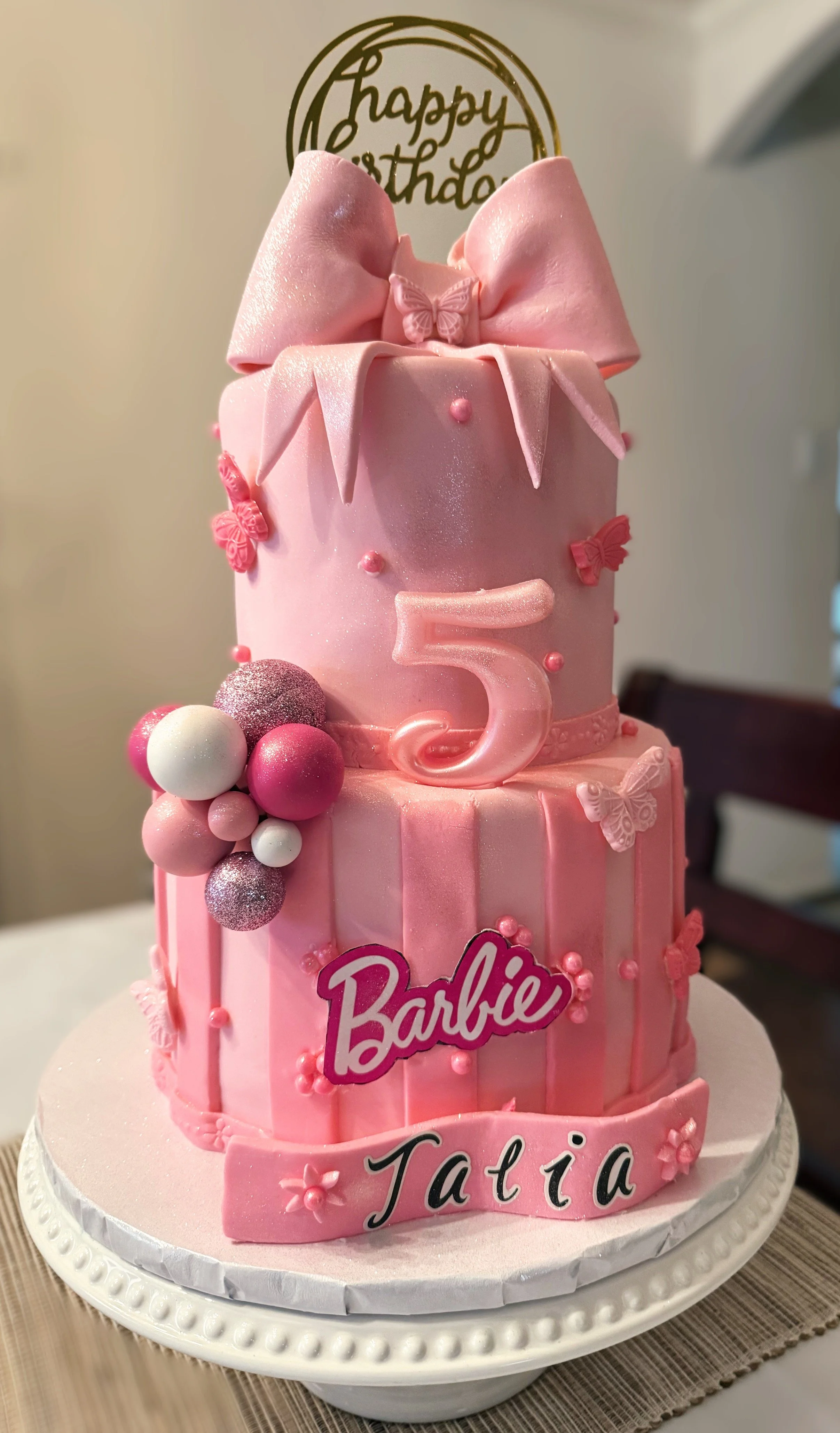 Pink three-tier Barbie-themed birthday cake decorated with bows, butterflies, and pink and white balls, with a 'Happy Birthday' topper and the name 'Tatía' on the bottom tier.