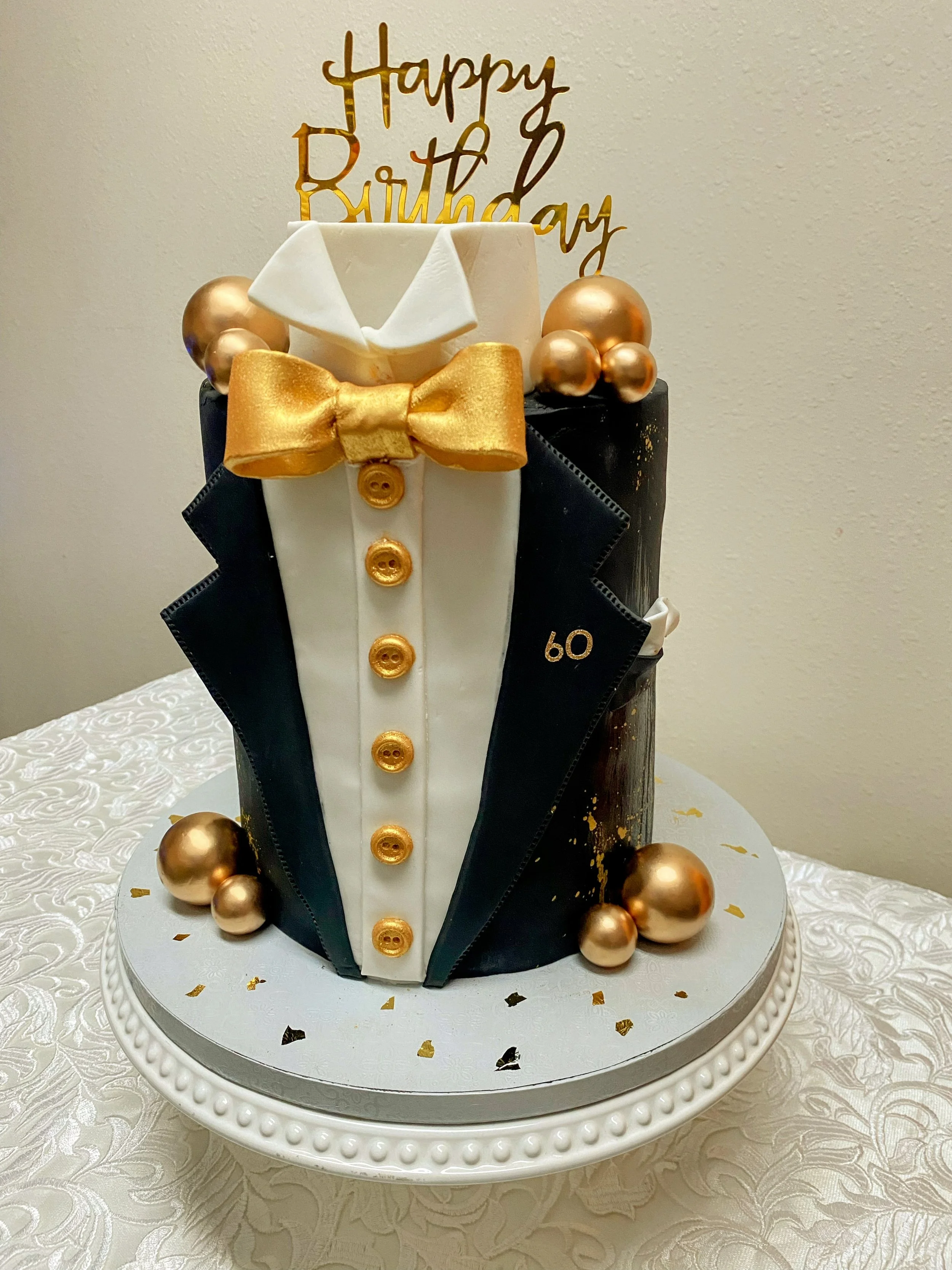A birthday cake decorated with a tuxedo design, gold buttons, and gold 'Happy Birthday' topper. The cake has gold and bronze balls around it and sits on a white textured tablecloth.