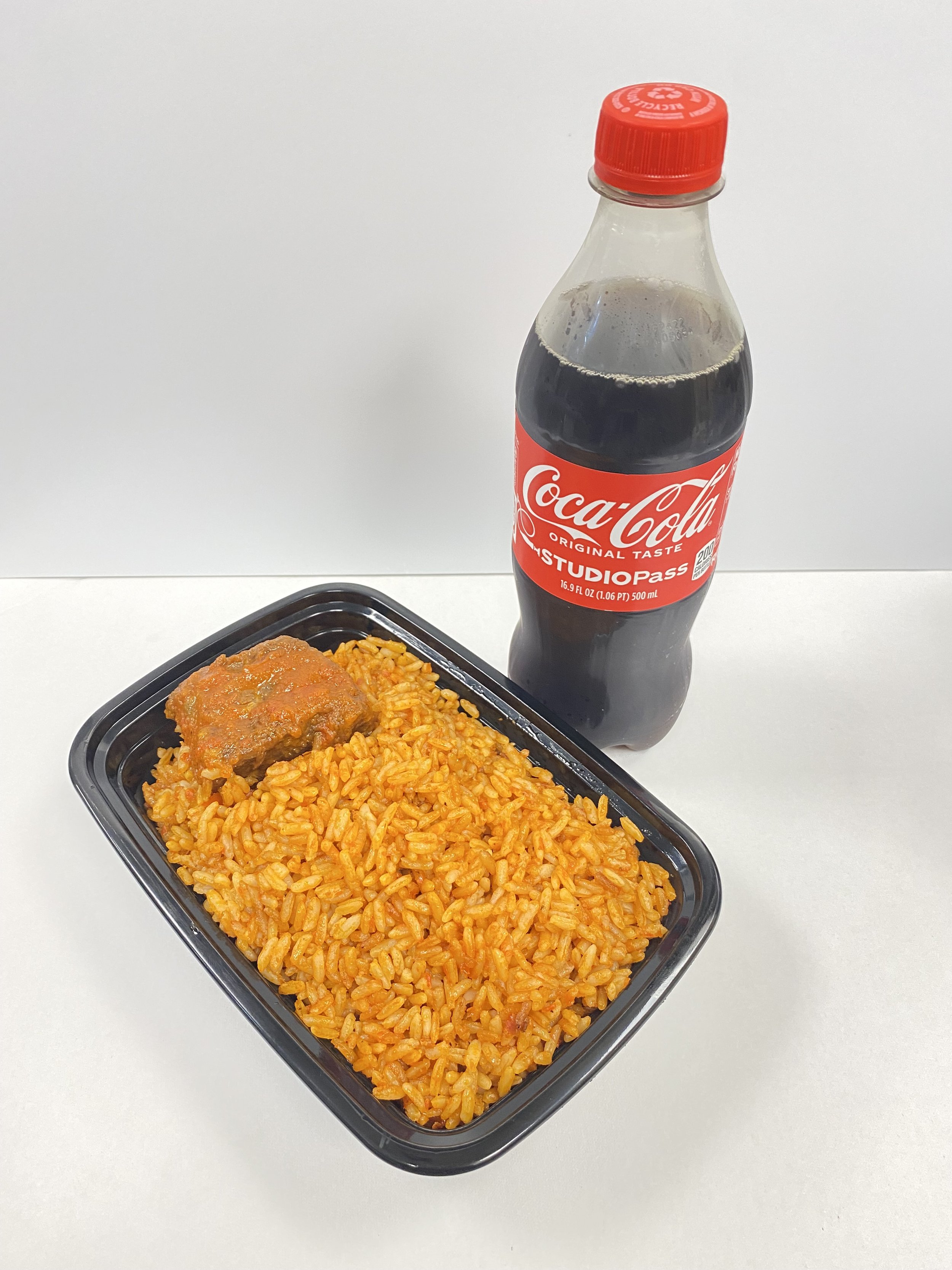 A black plastic tray filled with orange-colored rice and a meatball with sauce on top, placed on a white surface. A bottle of Coca-Cola is positioned behind the tray.
