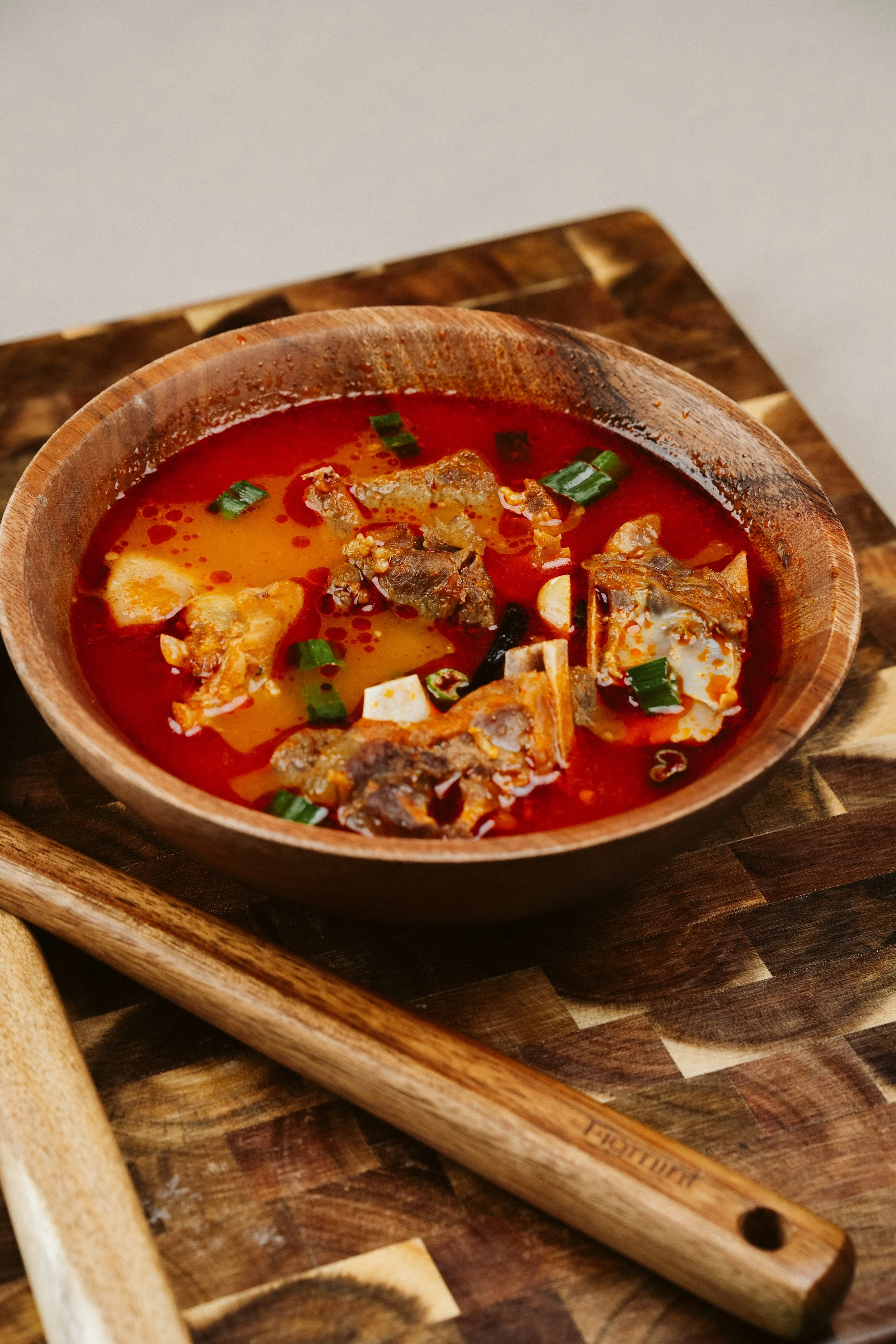 A bowl of spicy red soup with chunks of meat, tofu, and green onions on a wooden surface.