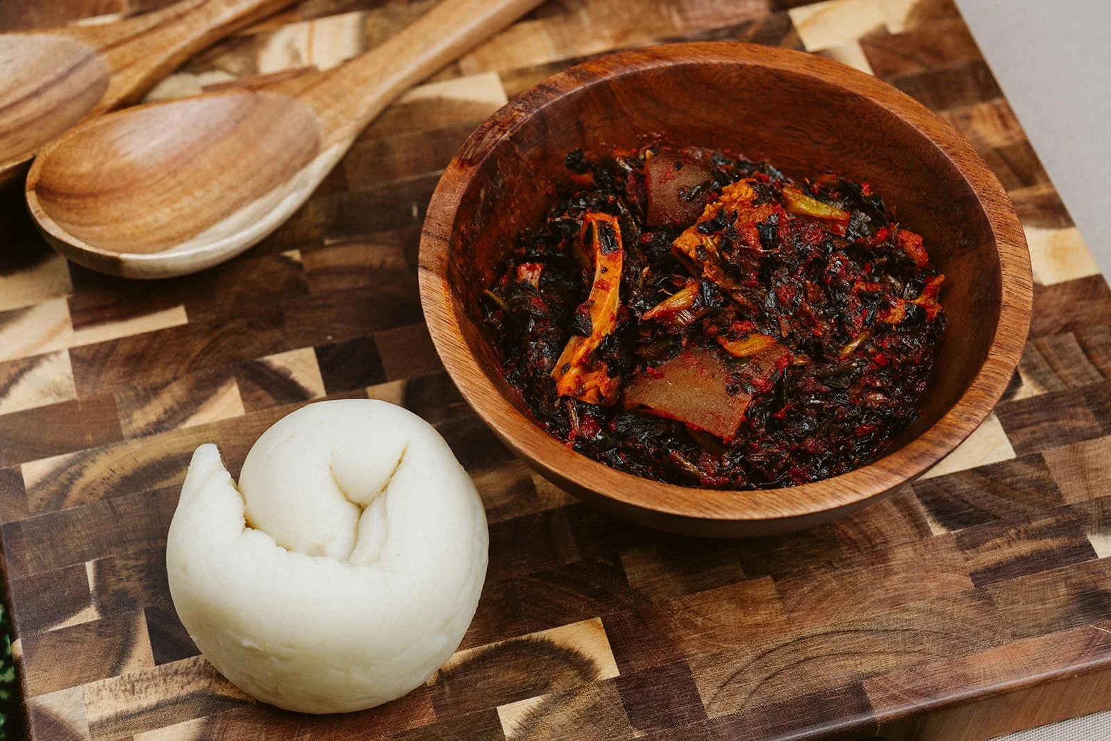 A wooden bowl filled with a spicy stew containing vegetables and meat, placed on a checked wood cutting board. Next to the bowl is a ball of mochi, and a wooden spoon rests on the board.