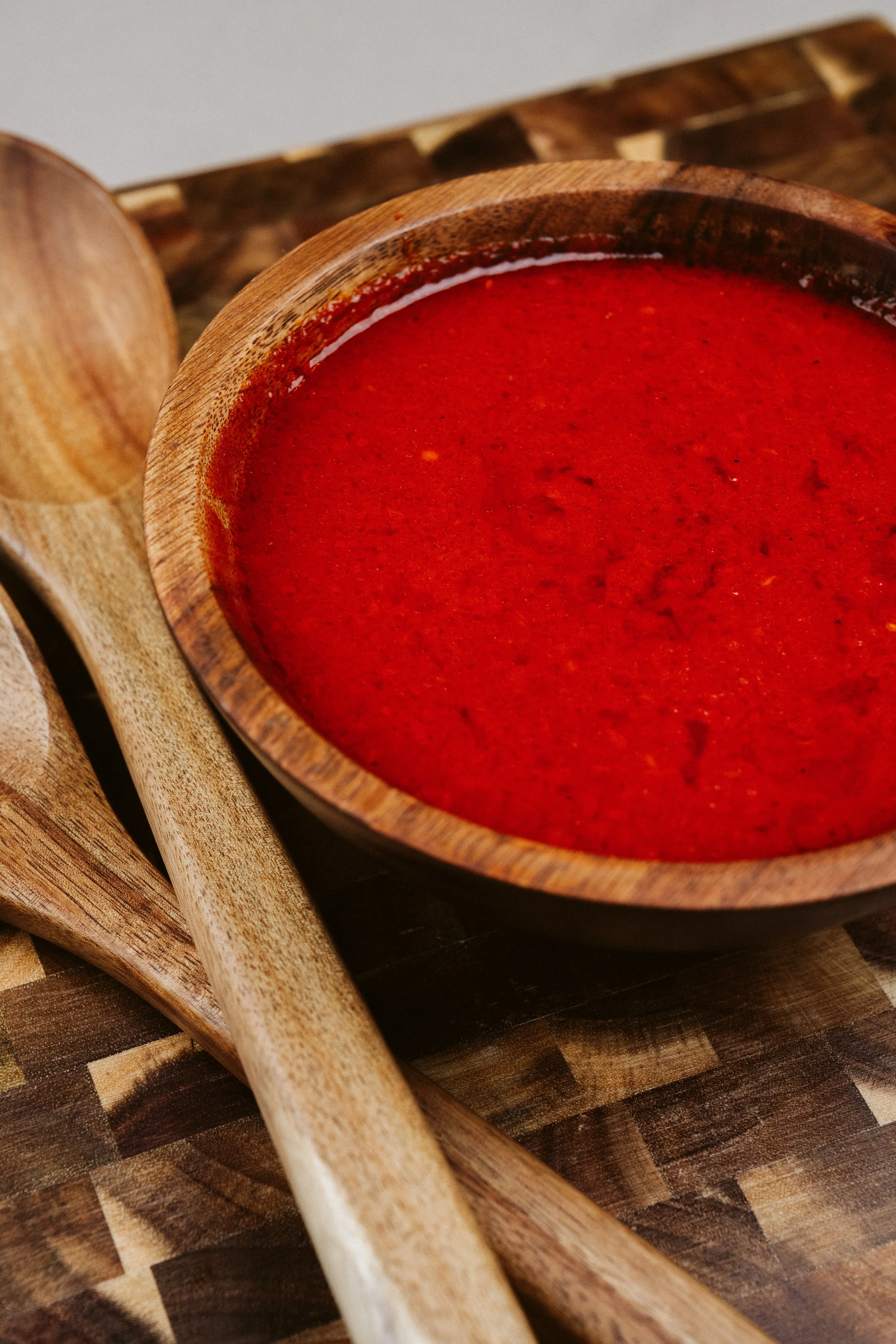Close-up of a bowl of red sauce or paste in a wooden bowl, with wooden utensils on a wooden surface.